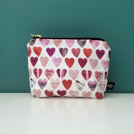 A Small Hearts wash bag placed on a white shelf with a blue green background. The wash bags has a gold metal zipper and a Brand label sewn into the outside seam.