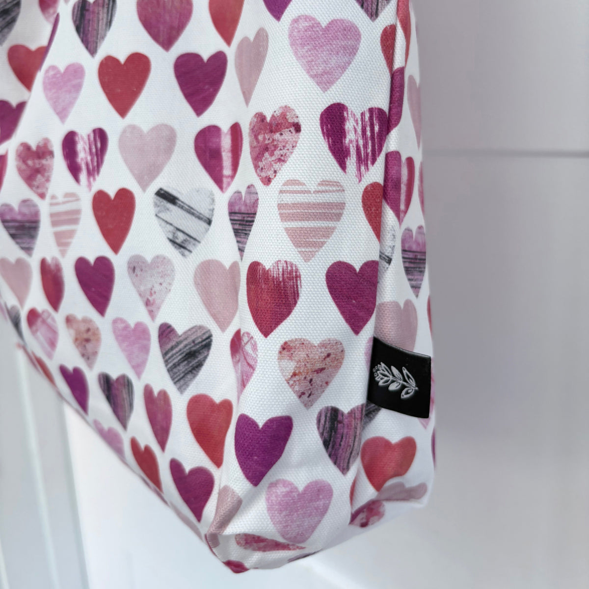 A white canvas tote bag featuring an all-over pattern of hearts in shades of pink, red, and purple, with a small black brand tag near the bottom right corner.