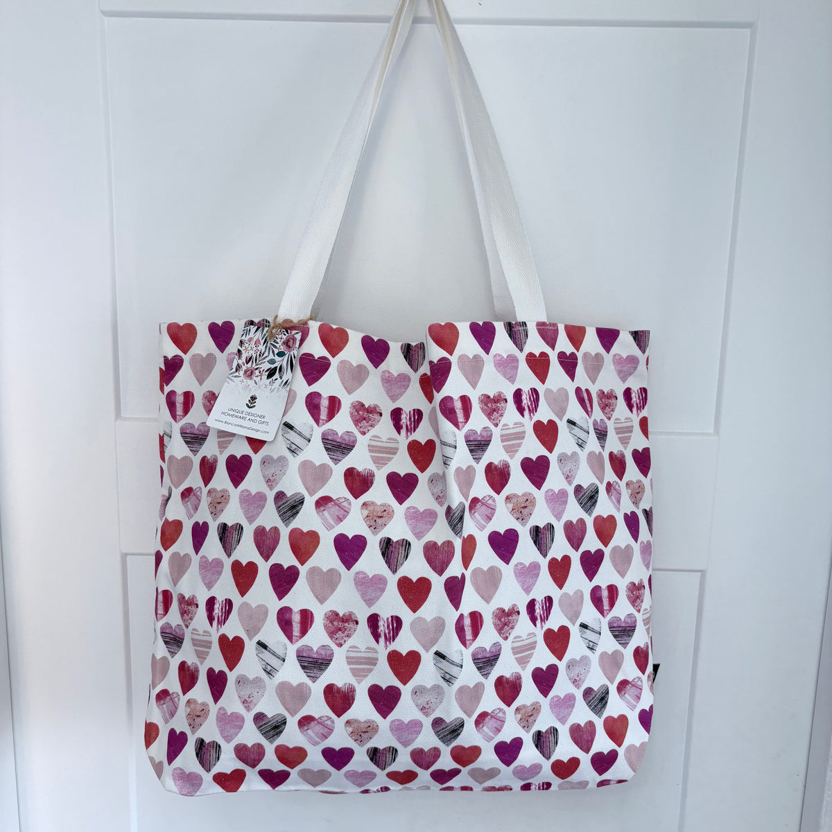 A white tote bag with a repeating pattern of red, pink, purple, and grey hearts, hanging from a metal hook on a white panelled door.