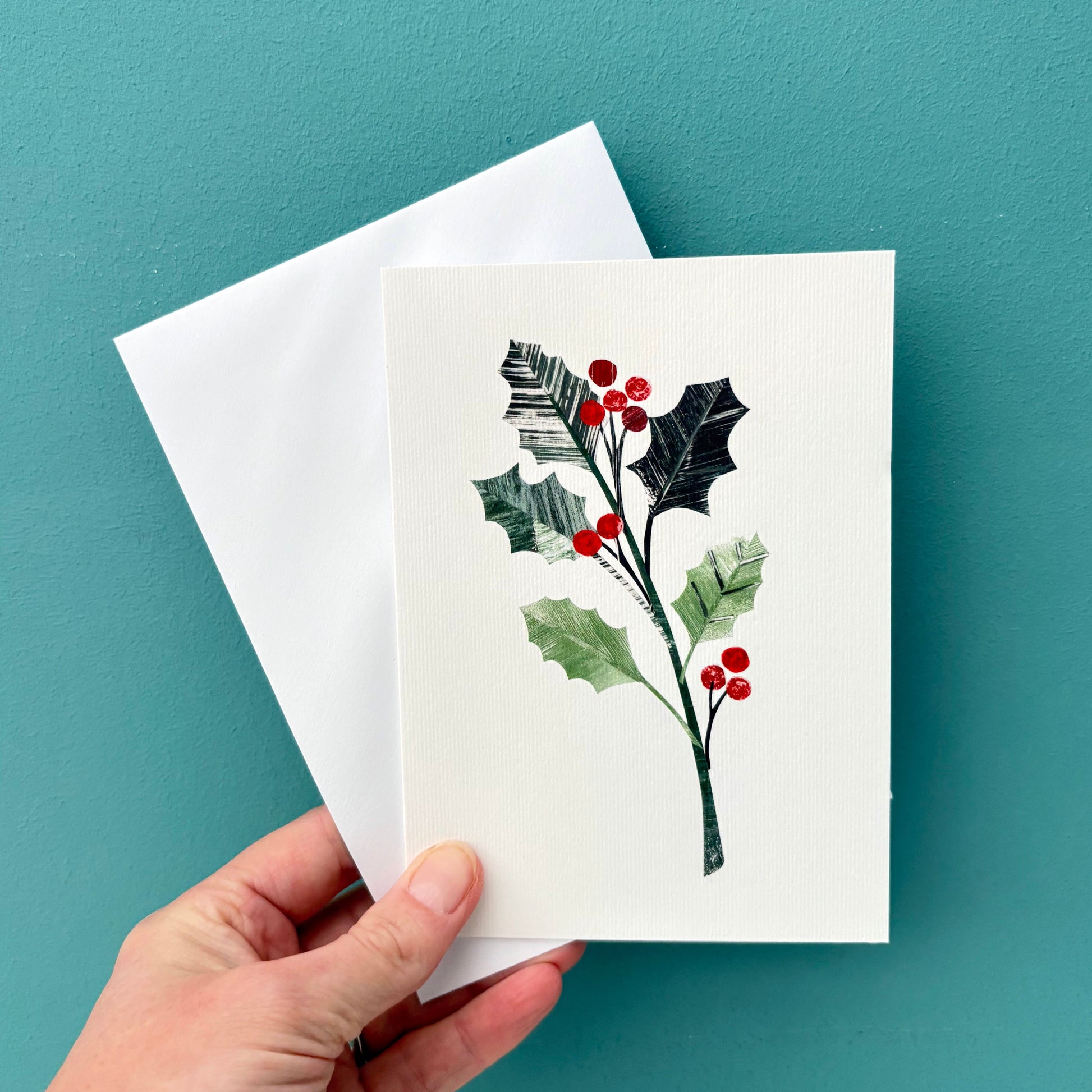 Hand holding a card with holly design and a white envelope against a teal background.