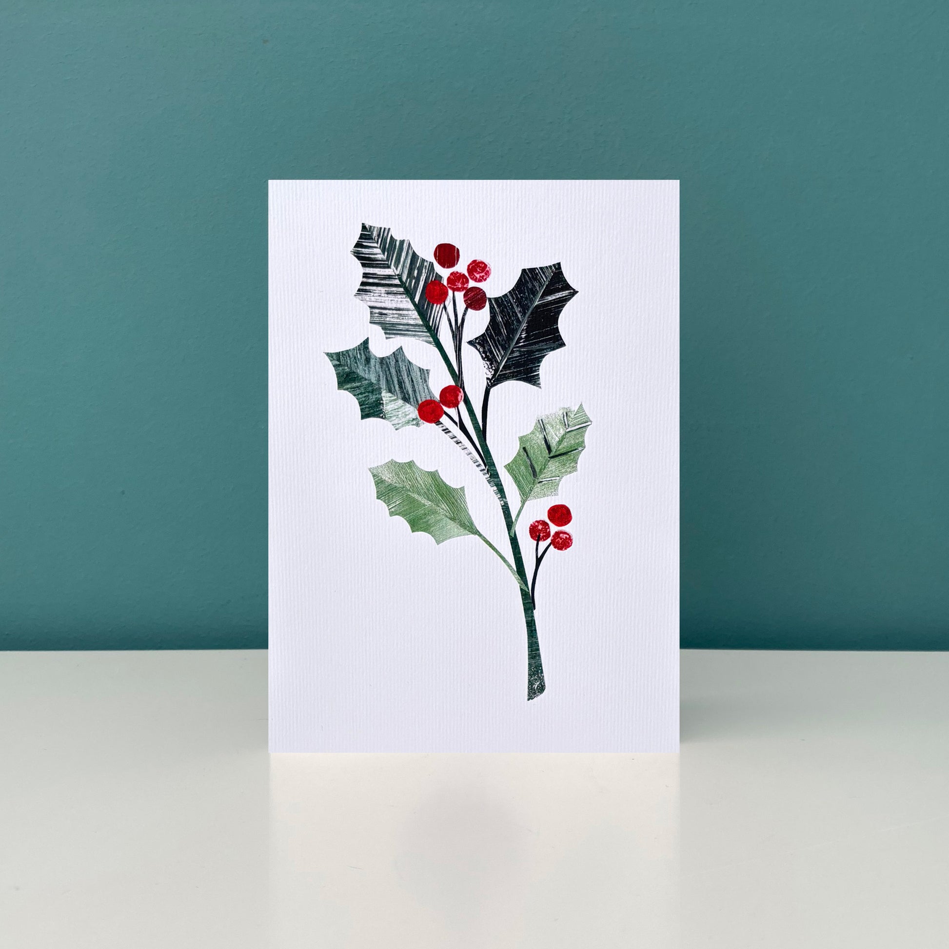 Greeting card with holly design on a white background has been stood up on a white shelf against a Teal wall.