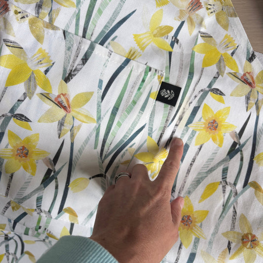 A close-up of a white fabric garment with a yellow daffodil and green leaf print, being held by a hand wearing a light blue ribbed sweater. A small black brand tag is visible on the fabric.