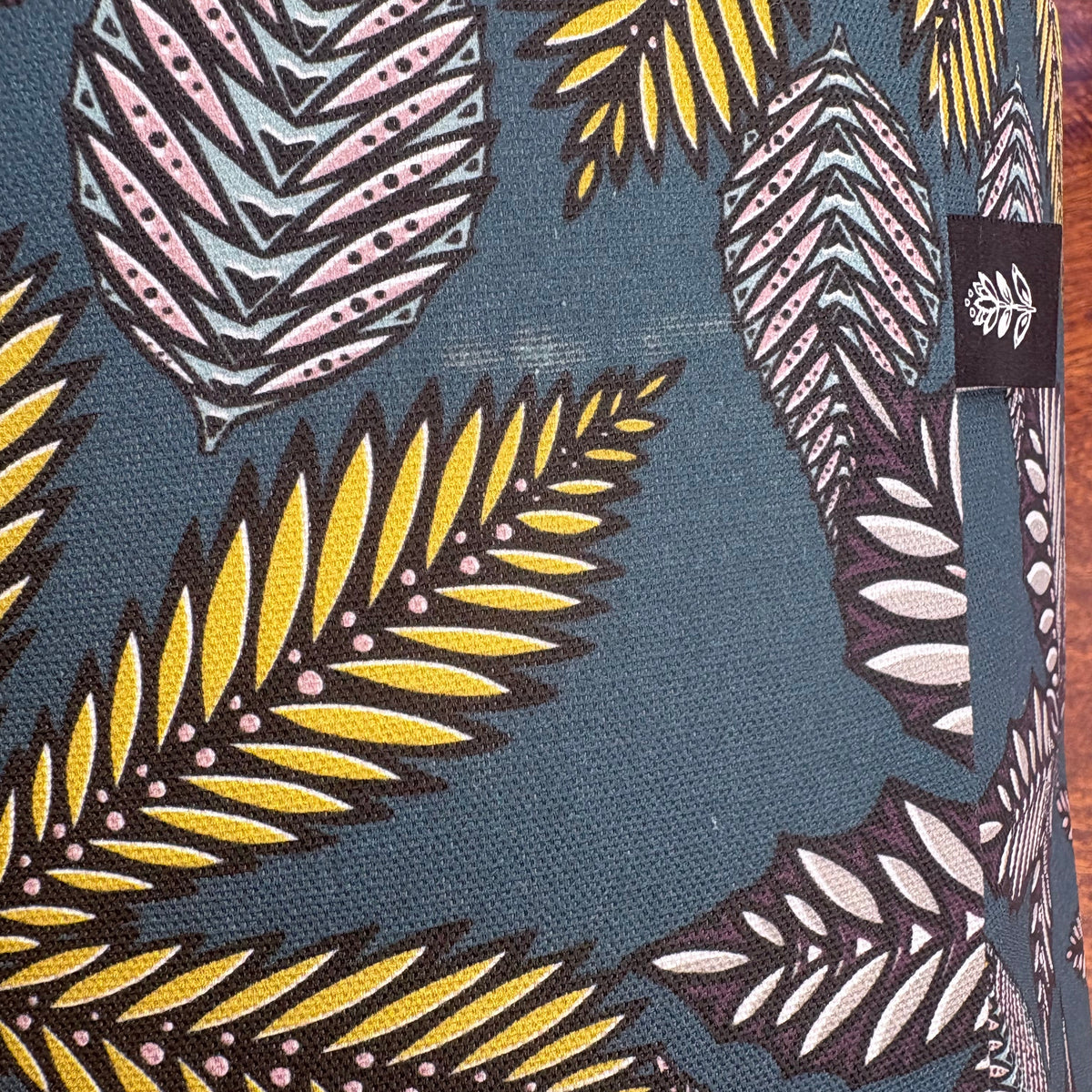 A close-up of a patterned fabric featuring stylized tropical leaves in yellow, pink, white, and gray on a dark teal-blue background. A small black fabric brand tag is visible on the right side.