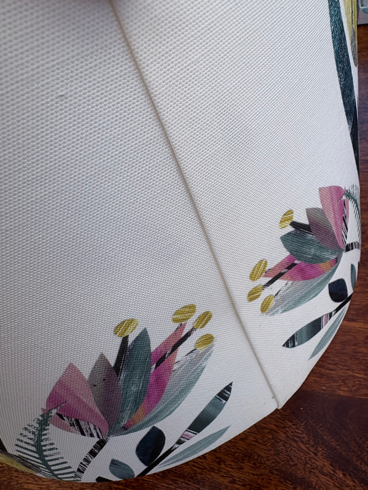 Close-up of a white woven fabric lampshade, featuring a botanical pattern with pink, green, and gold accents, resting on a dark wood floor.