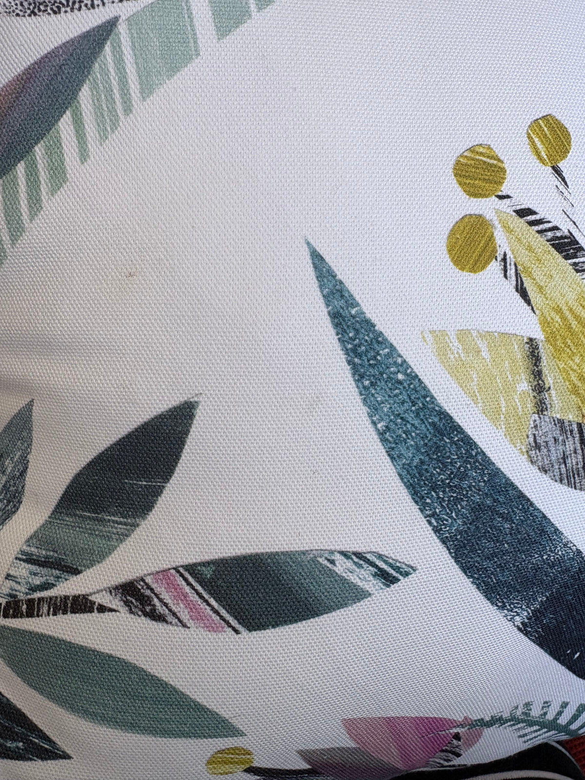 A close-up of a fabric surface with an abstract botanical print on a white base, featuring teal, blue-green, yellow, pink, and gray brushstroke-style leaves and circular floral elements.
