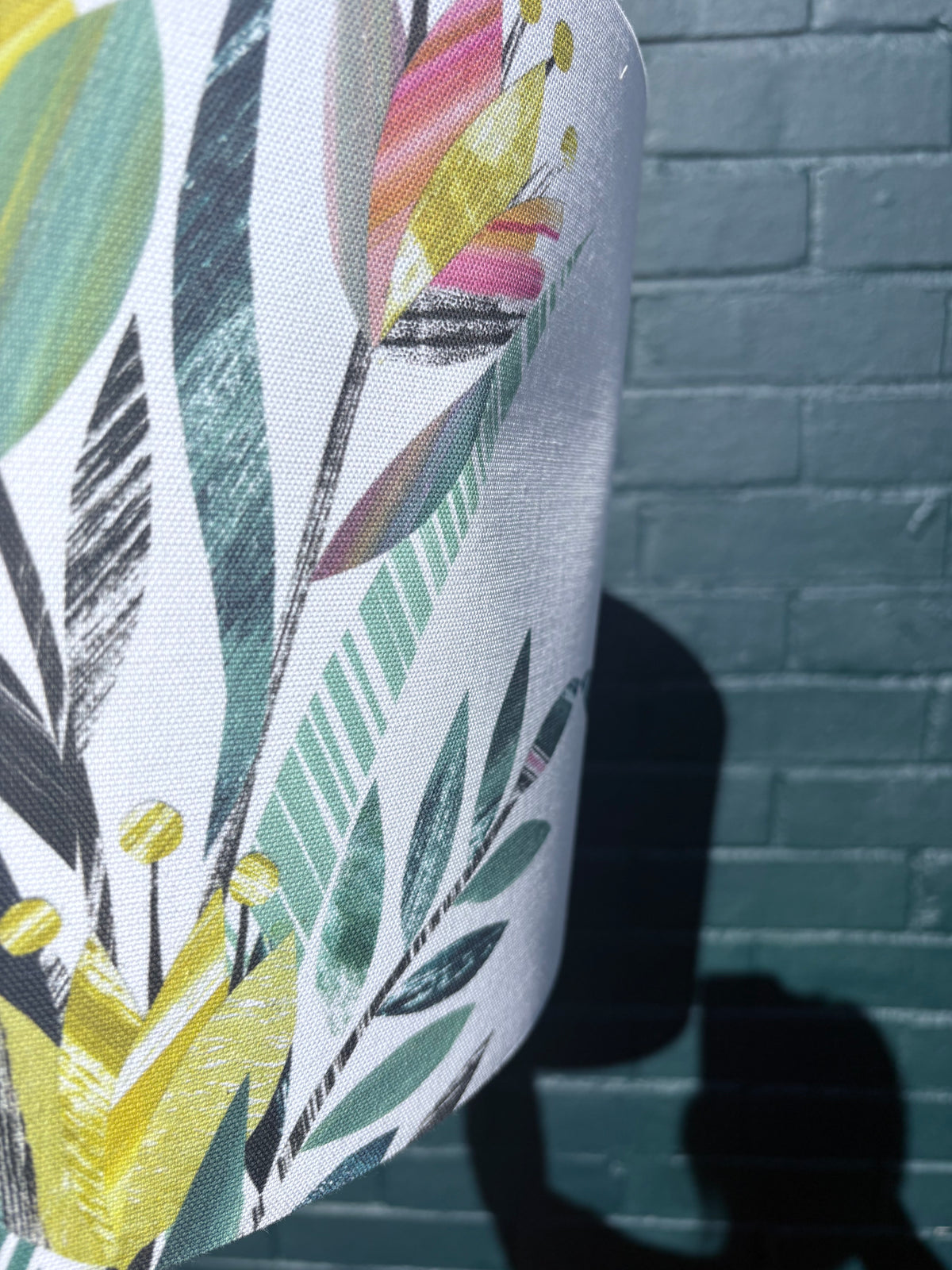 A close-up of a white fabric lampshade featuring a colourful tulip floral and leaf pattern, shown against a dark teal brick wall with a strong shadow cast on the right.