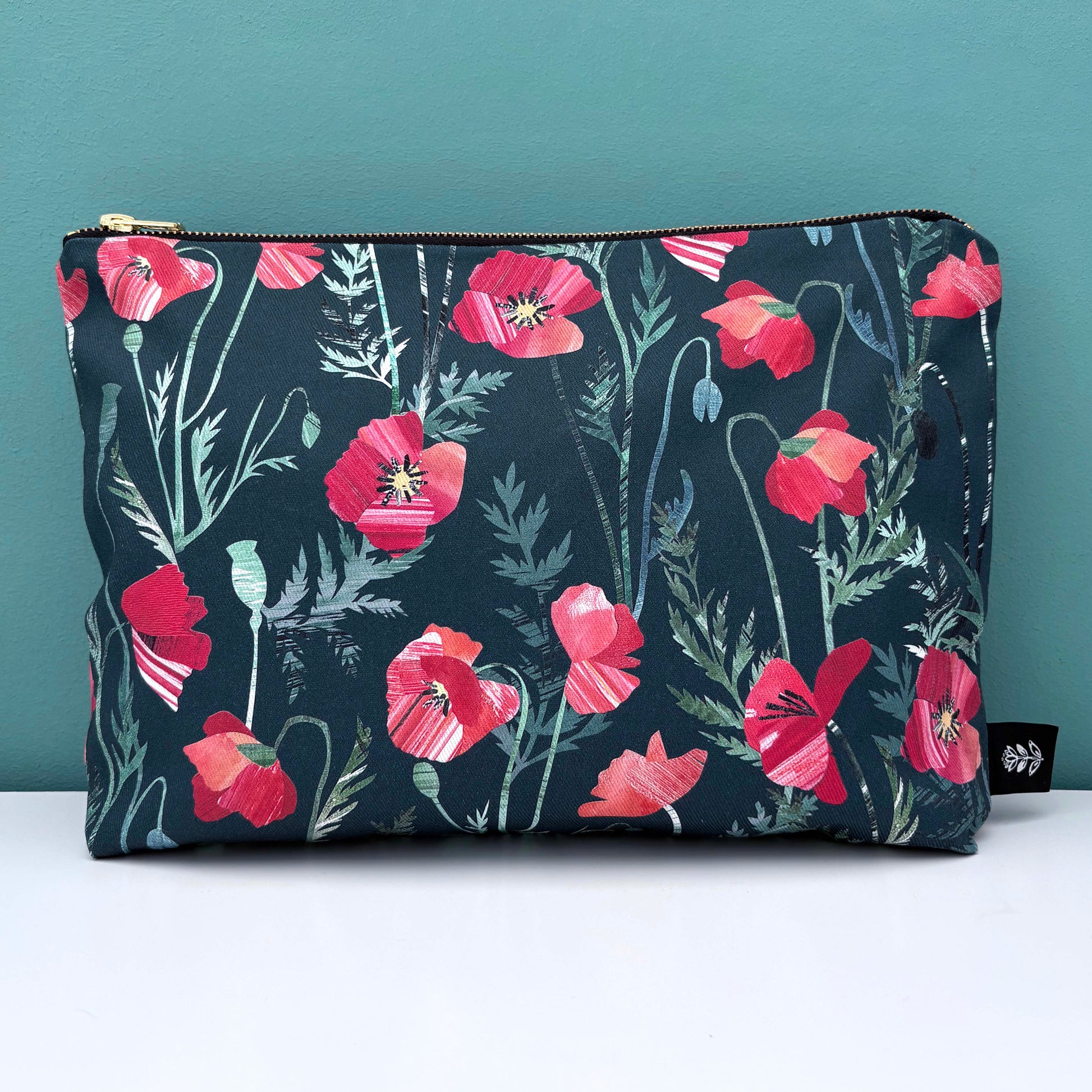 A large Poppy wash bag featuring pretty red poppies with green leaves on a teal background has been placed on a white shelf with a blue green wall behind it.