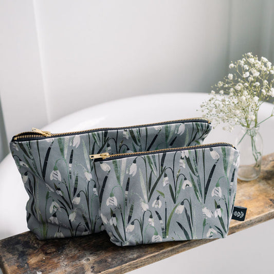 Two Snowdrop floral-patterned wash bags are on a wooden surface with a candle and flowers in the background.