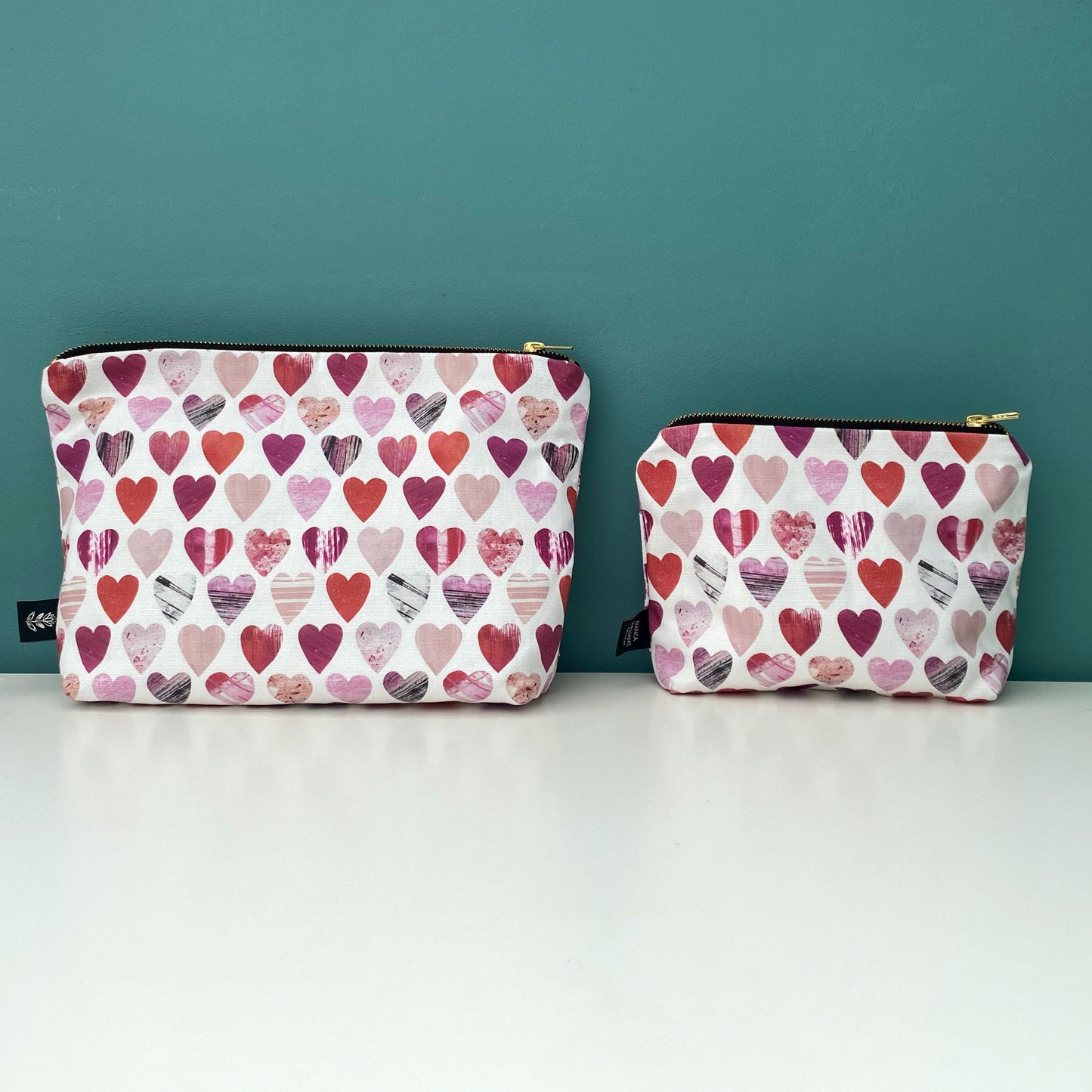 A large and small Hearts Wash Bag have been placed side by side on a white shelf.  They have gold metal zip and a Brande label sewn into the outside seam.