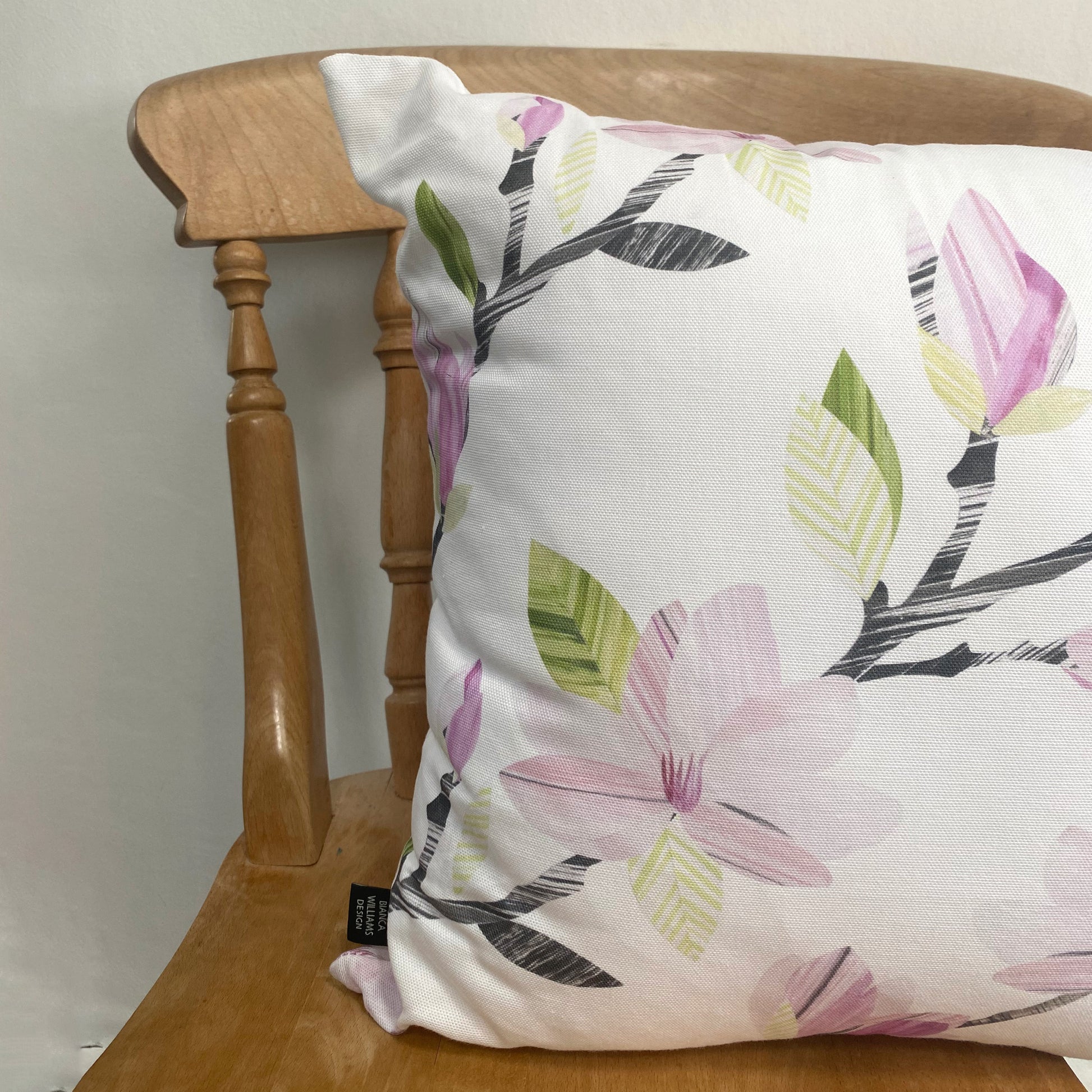 A square magnolia Cushion has been placed on a wooden chair against a pale grey wall.