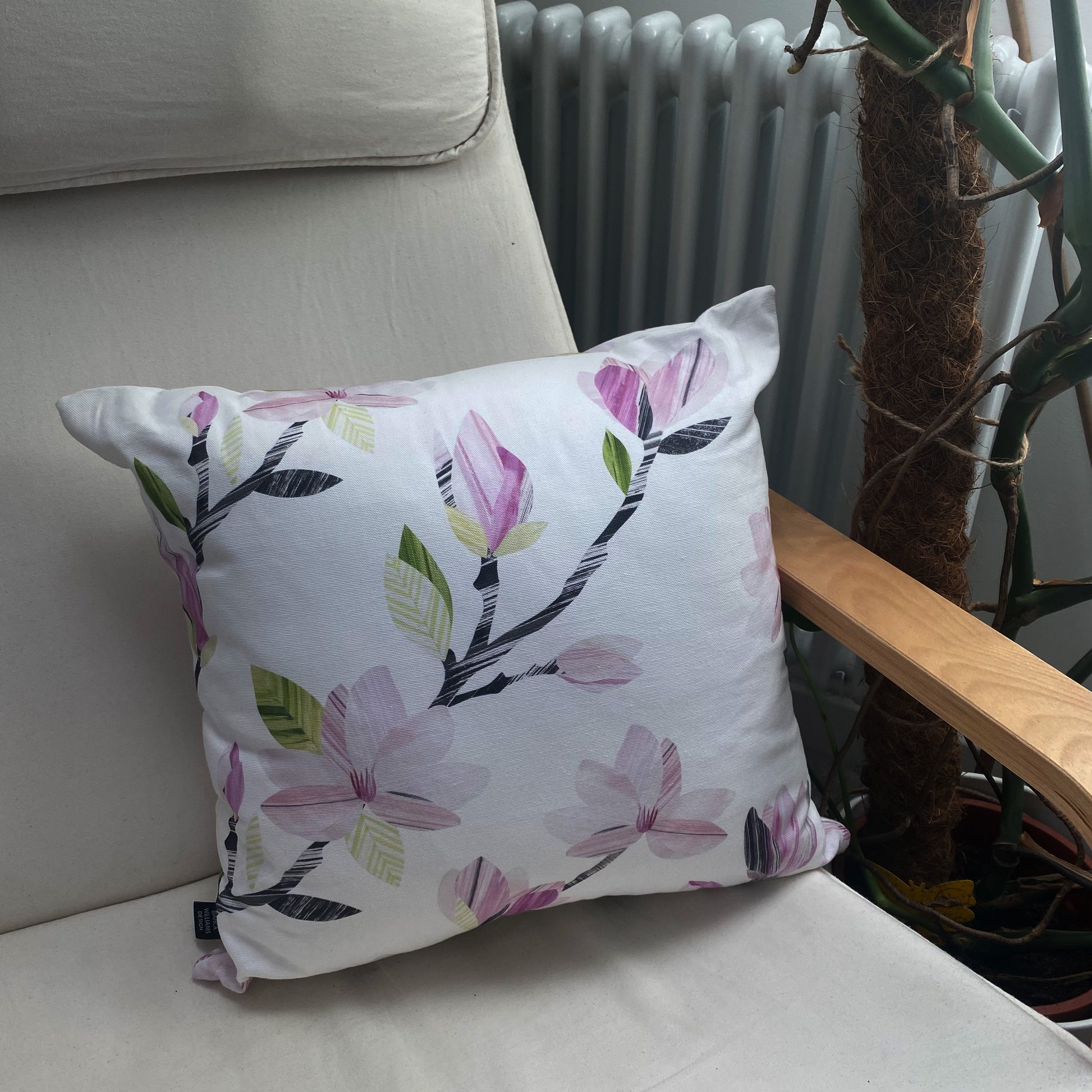 A Magnolia Cushion has been placed on a cream canvas and wooden arm chair.  A radiator and plant can be seen in the background.