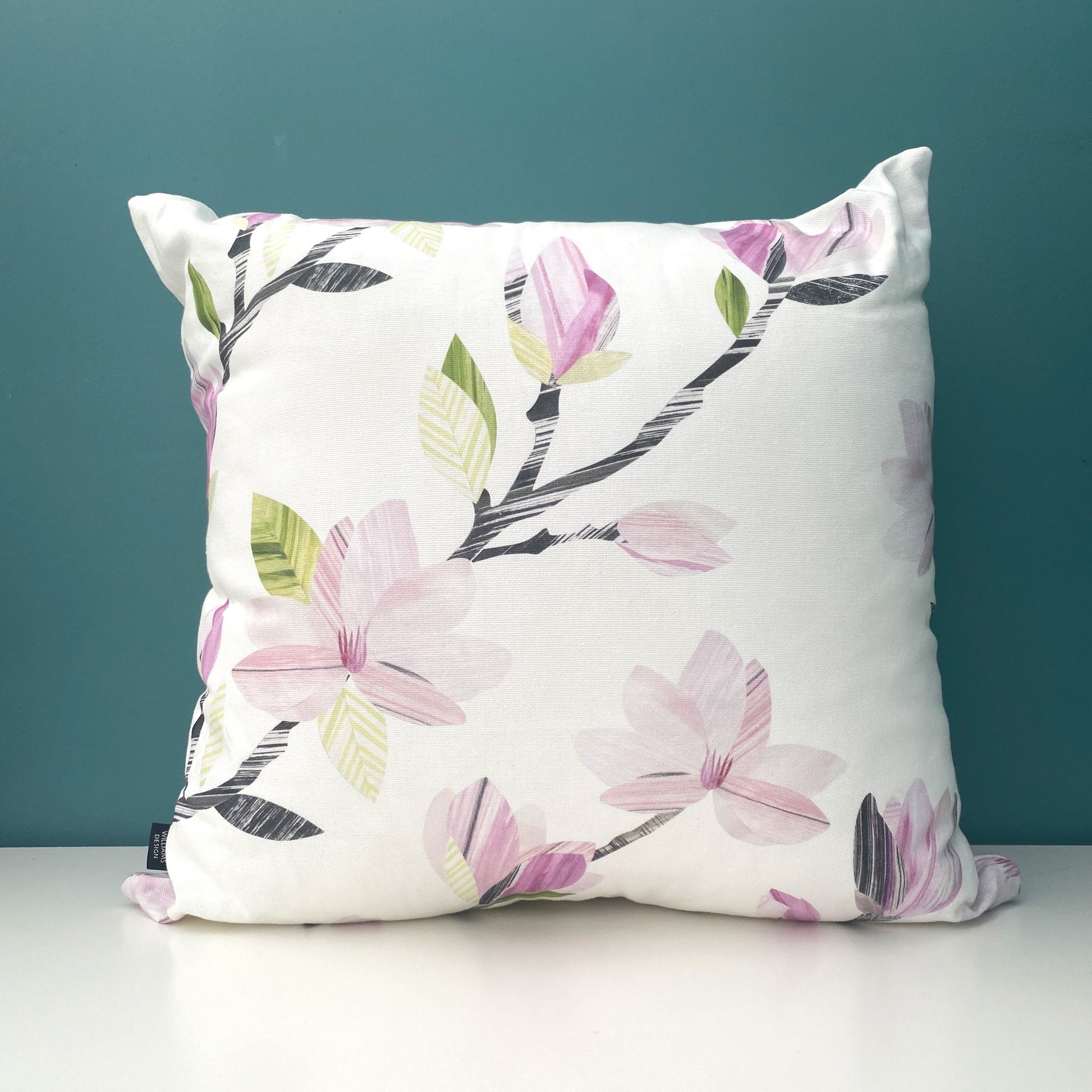 A Square Magnolia Cushion, featuring soft Pink Magnolia Flowers with grey and green leaves on a white background has been placed on a white shelf against a blue/green wall.