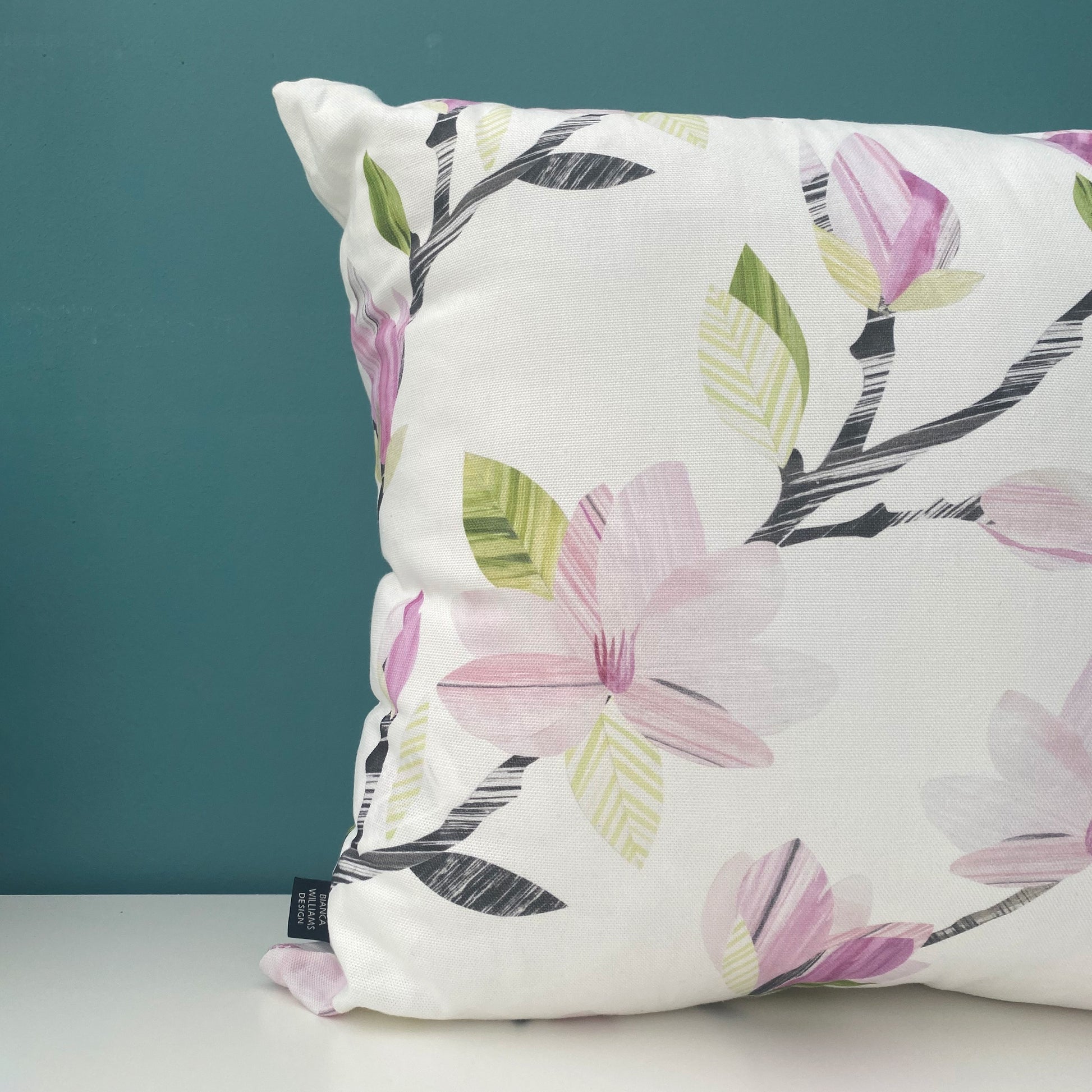 A close up of a Square Magnolia Cushion, featuring soft Pink Magnolia Flowers with grey and green leaves on a white background has been placed on a white shelf against a blue/green wall.