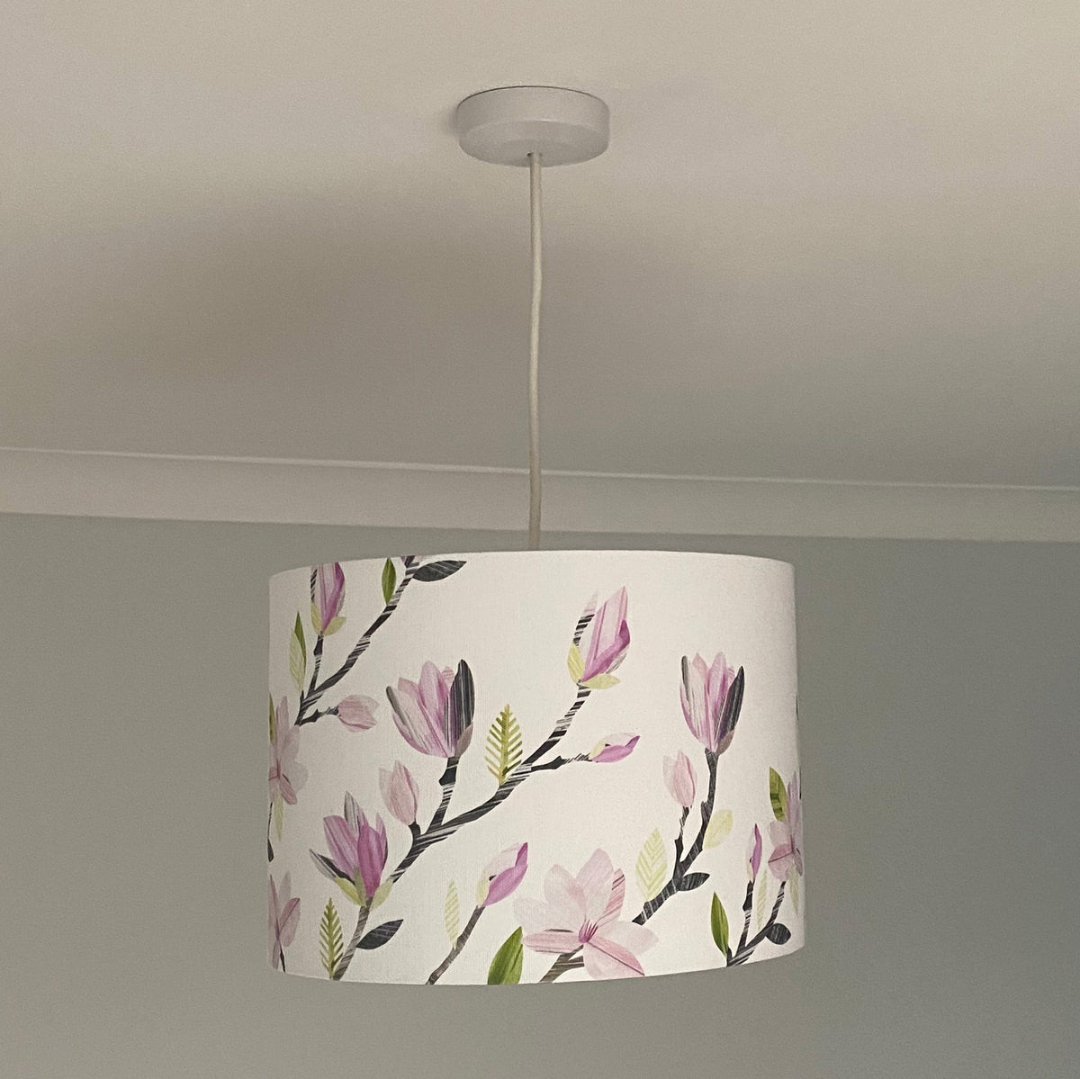 A white drum pendant light shade featuring a watercolor-style floral print of pink magnolia blossoms and green leaves, hanging from a white ceiling.