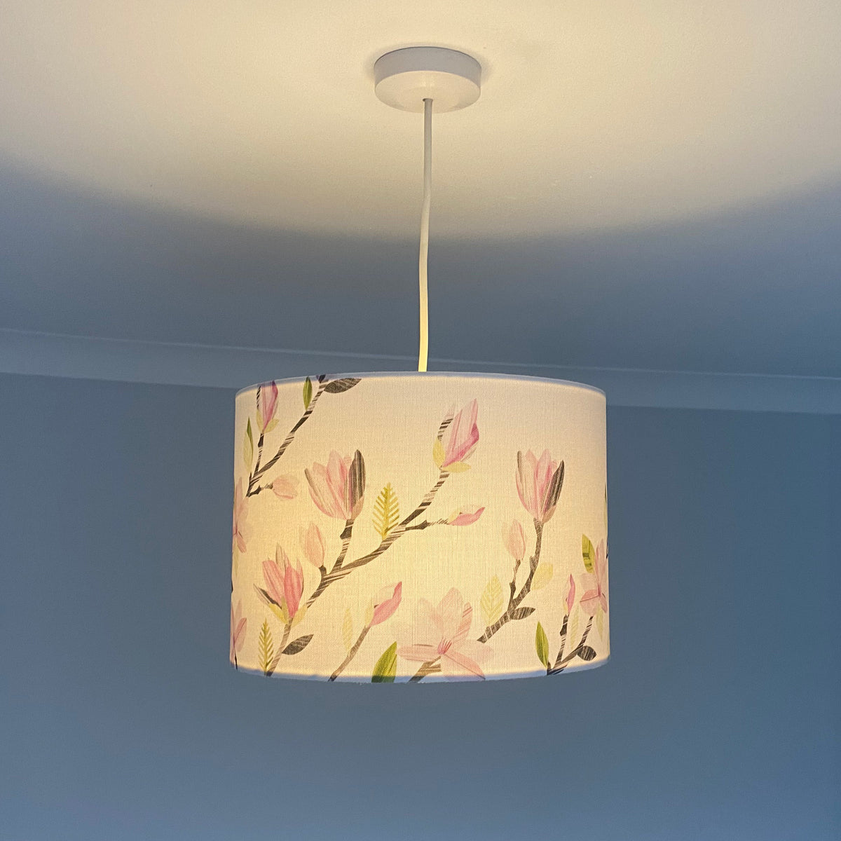 A drum pendant lamp with a cream-colored fabric shade featuring a pink and green magnolia floral pattern, hanging from a white ceiling canopy against a blue-gray wall.