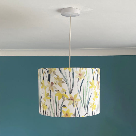 Daffodil Lampshade with a pendant ceiling fitting, in medium size.  Has Yellow Daffodil flowers with Green Leaves on a plain white background.  The lampshade is photographed against a blue/green background.