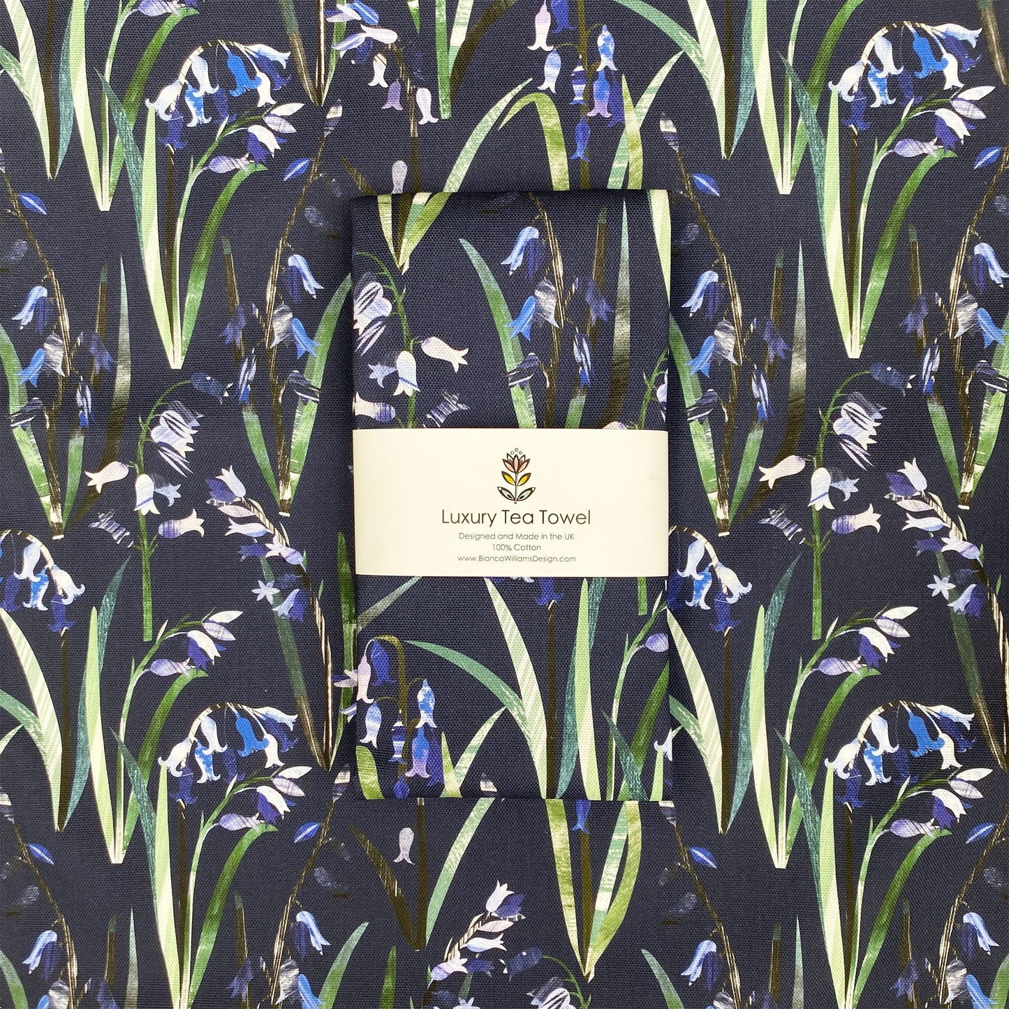 A navy Bluebell tea towel has been packaged in a branded belly band and has been placed on an open out tea towel in the same colour and design.