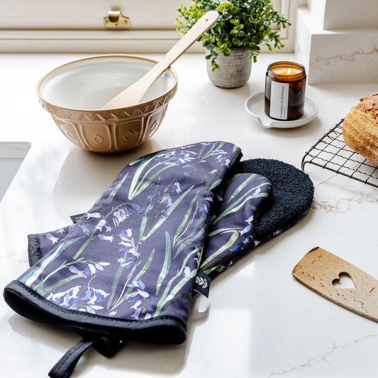 A pair of oven mitts with a dark purple and green Bluebell botanical pattern and black trim, resting on a white marble kitchen counter next to a ceramic bowl, a small plant, and a candle.