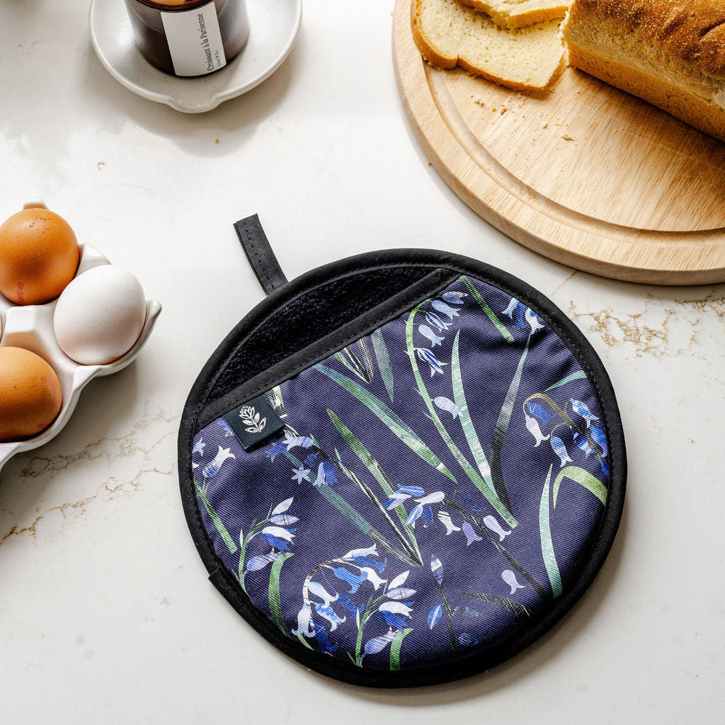 A round potholder with a navy blue background and a blue-and-green bluebell floral print, styled on a light marble surface with bread, eggs, a candle, and a plant nearby.