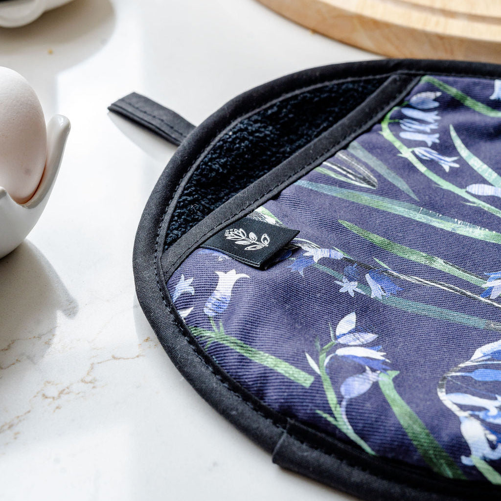 A close-up of a round potholder with a navy blue background, featuring bluebells, shown on a light countertop with a wooden cutting board, bread, and a small dish in the background.
