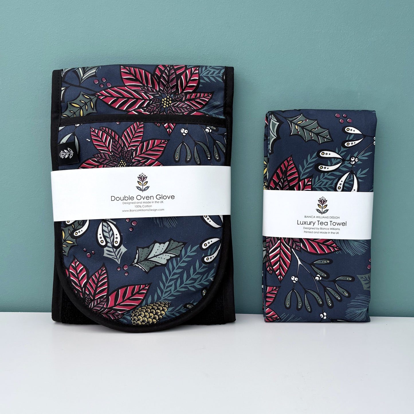 A pair of Navy Poinsettia Oven Gloves and matching tea towel have both been packaged in a branded belly band and  placed on a white shelf against a blue green background.