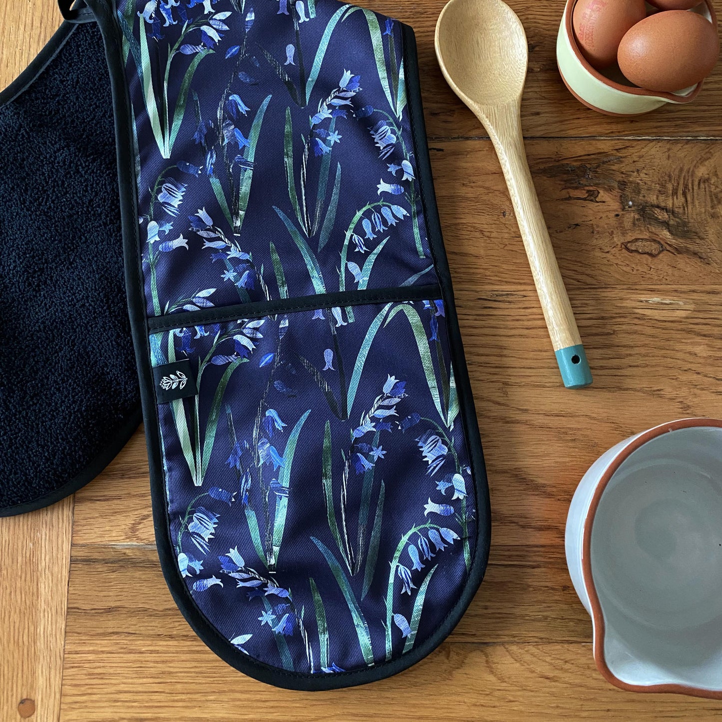 Navy Bluebell Oven Gloves placed on a wooden table at an angle so that the black towelling backing can be seen.  