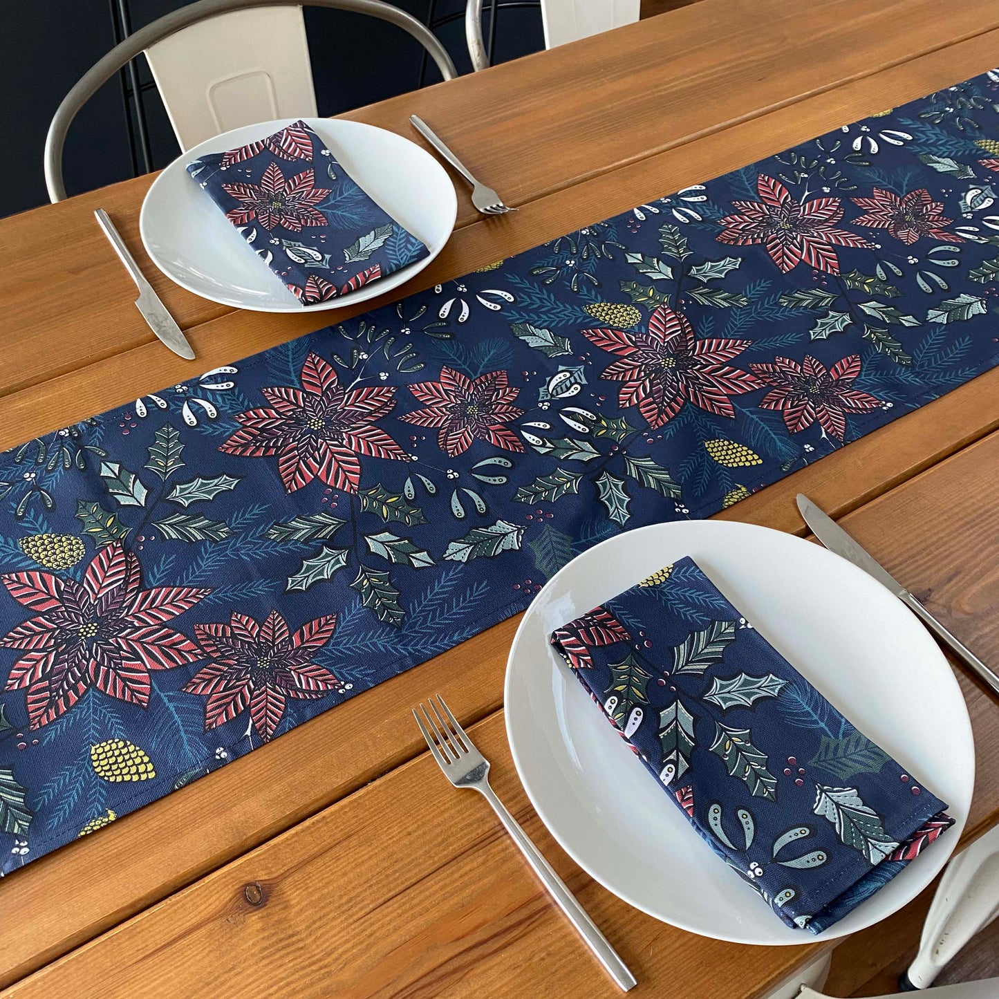 A Christmas Table Runner featuring Red Poinsettia flowers and green holly on a Navy background has been placed on a wooden Table, which has been set for two and two matching Napkins can  be seen on the plates.