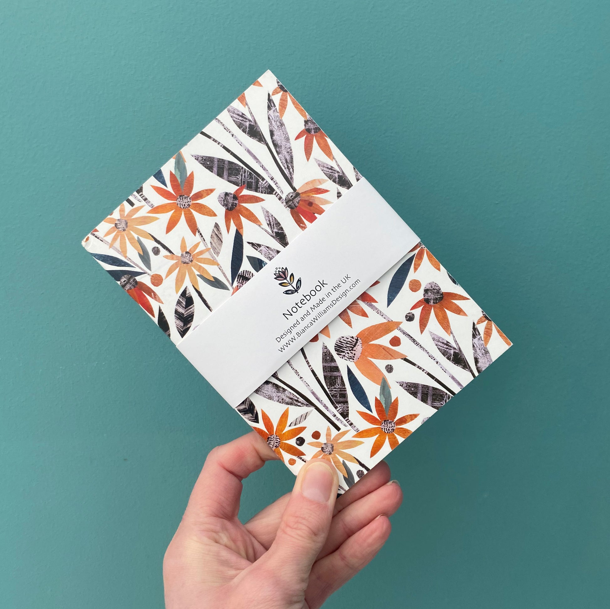 Small A6 notebook featuring orange all over Coneflower patterns with green and grey leaves on a white background, packaged in a white branded belly band held in front of a blue green backdrop.