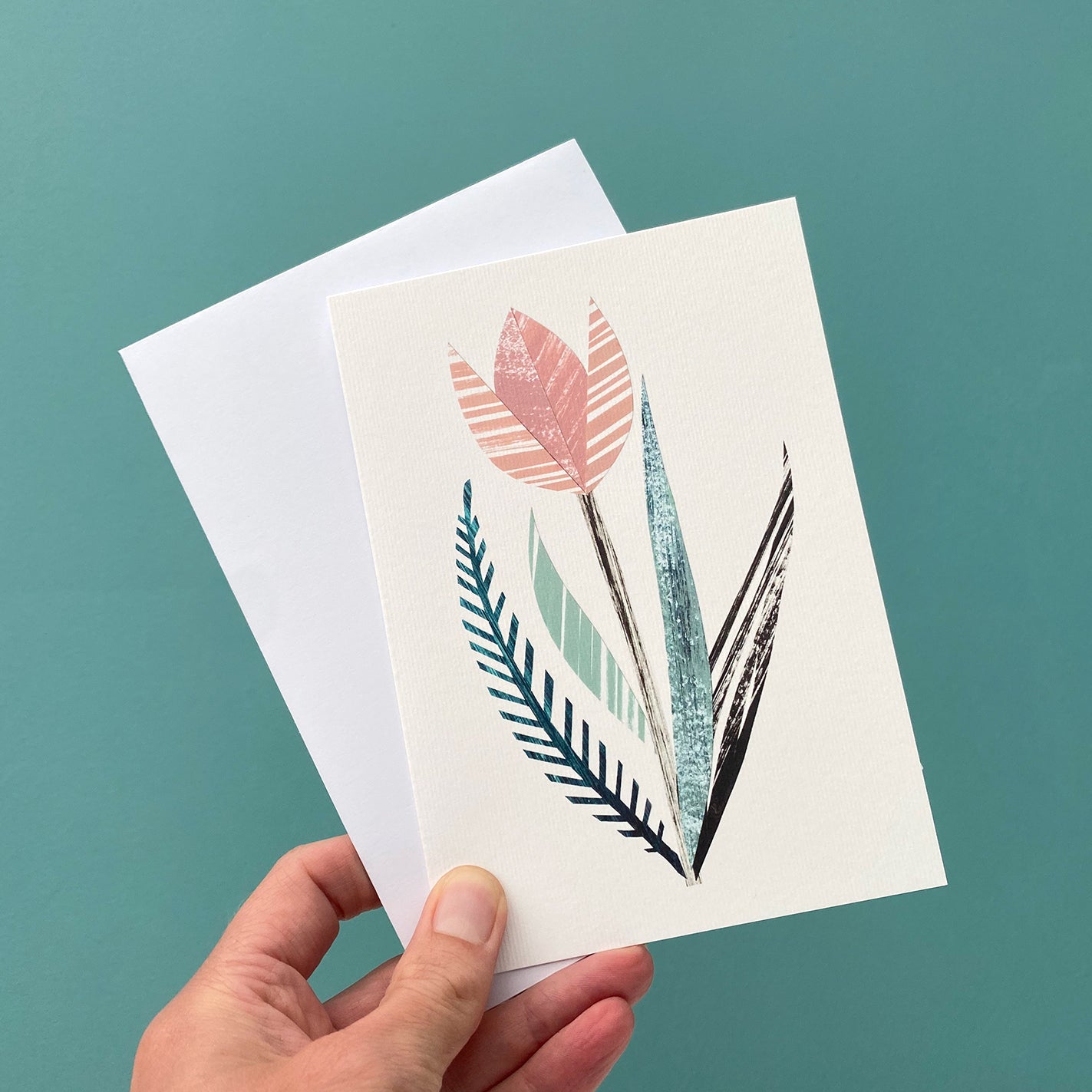 Card featuring a print of a stylised pink tulip with green leaves is being held with a white envelope against a blue green background.