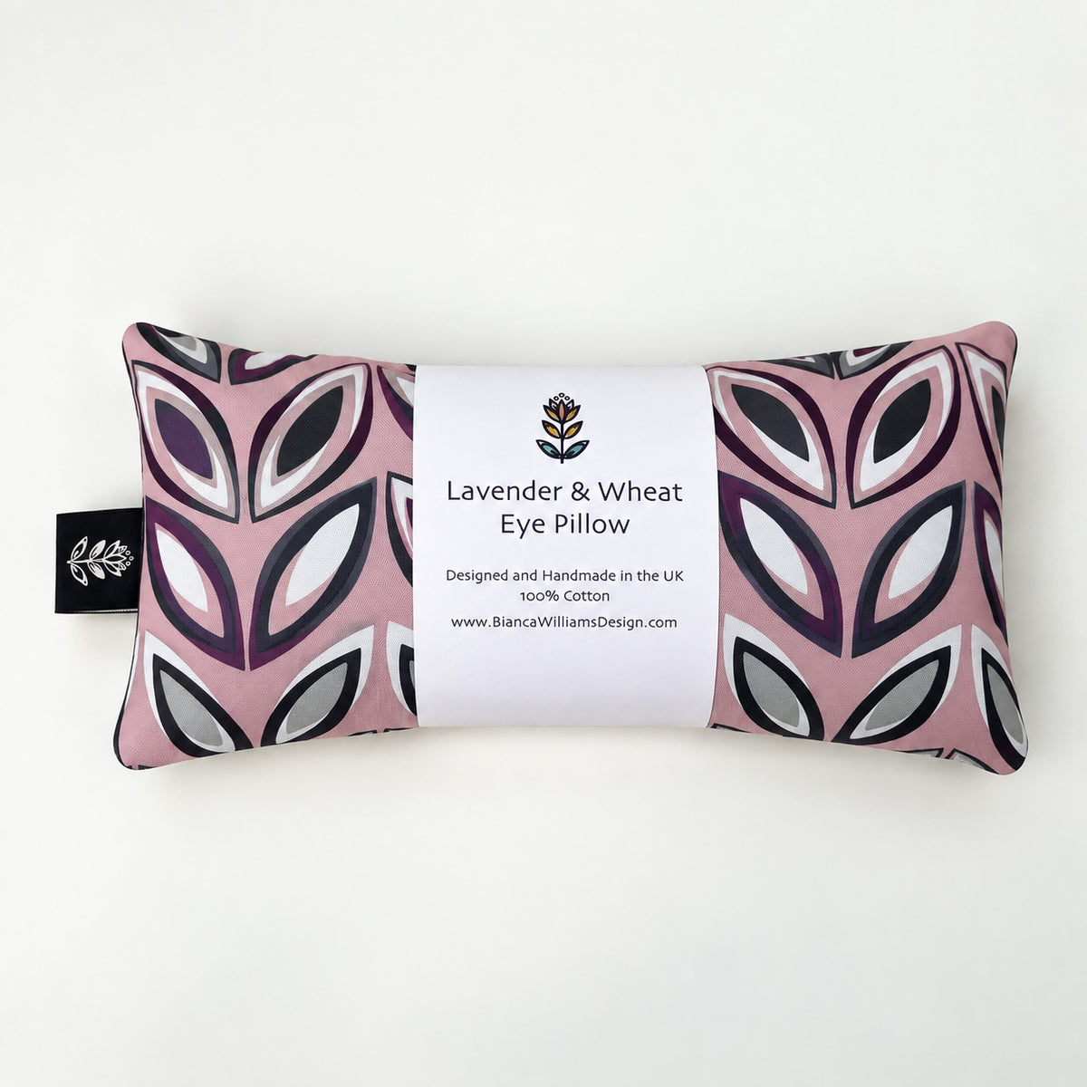 A rectangular lavender and wheat eye pillow by Bianca Williams Design, featuring a dusty pink, white, and dark purple geometric leaf pattern, wrapped in a white paper label.