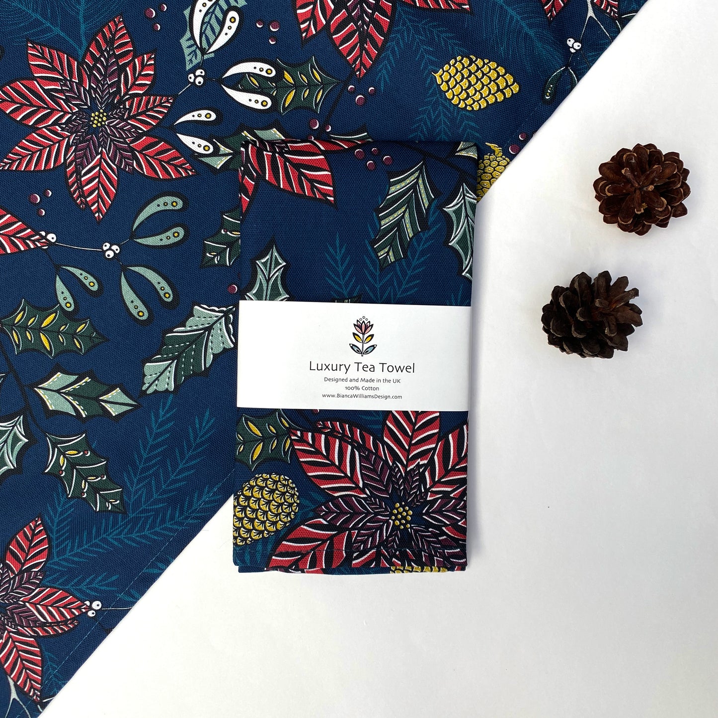 A Navy Poinsettia Tea Towel has been packaged in a Branded belly band and placed on a white background with a tea towel of the same design which has also been placed at an angle.