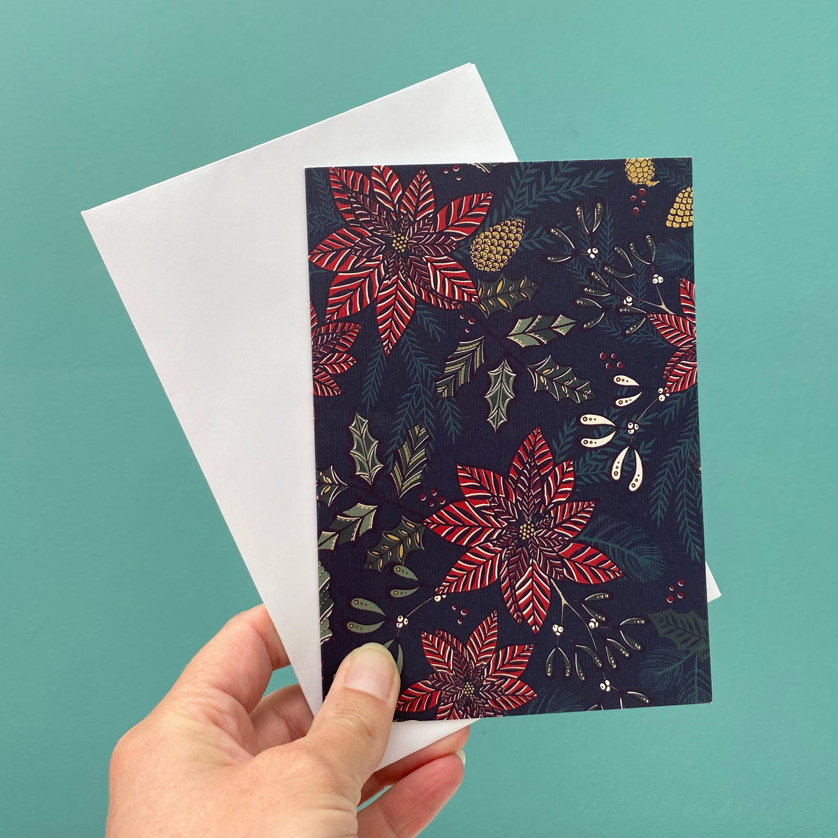 A hand holds a Christmas greeting card with a dark navy background and red poinsettia floral pattern, layered over a plain white envelope against a solid teal background.
