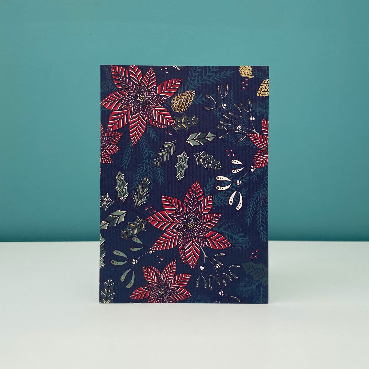 A vertical rectangular card standing upright on a white surface against a teal background, featuring a dark navy holiday floral print with red poinsettias, green leaves, and small gold pinecone motifs.