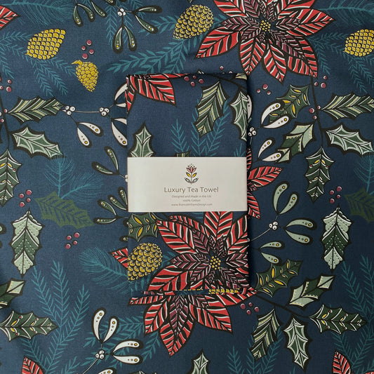 A Navy Poinsettia tea towel has been packaged in a branded belly band and has been placed on an opened tea towel of the same design.