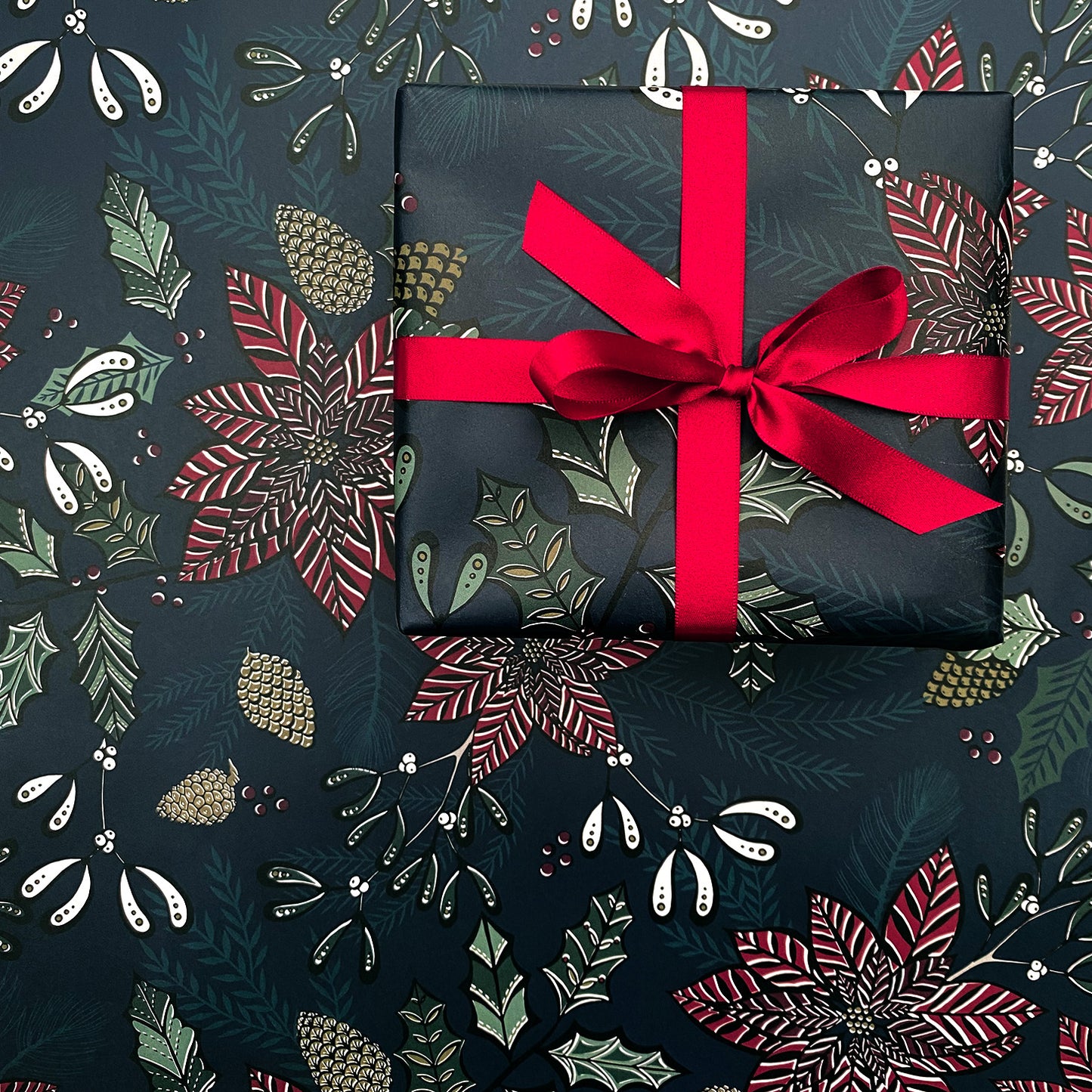 A present has been wrapped in the Navy Poinsettia Gift wrap and it has been tied with a matching red satin ribbon in a bow.  The present has been placed on a sheet of navy Poinsettia Gift wrap.