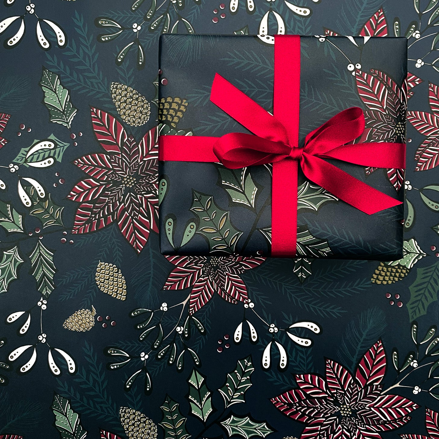 A present has been wrapped in the Navy Poinsettia Gift wrap and it has been tied with a matching red satin ribbon in a bow.  The present has been placed on a sheet of navy Poinsettia Gift wrap.