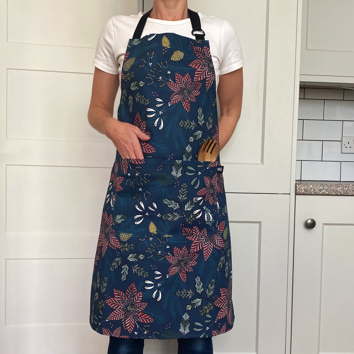 A lady is wearing the navy Poinsettia Christmas Apron which features red Poinsettia Flowers, with green holly and pine Cones.  The Apron has two front pockets and a black adjustable neck strap.