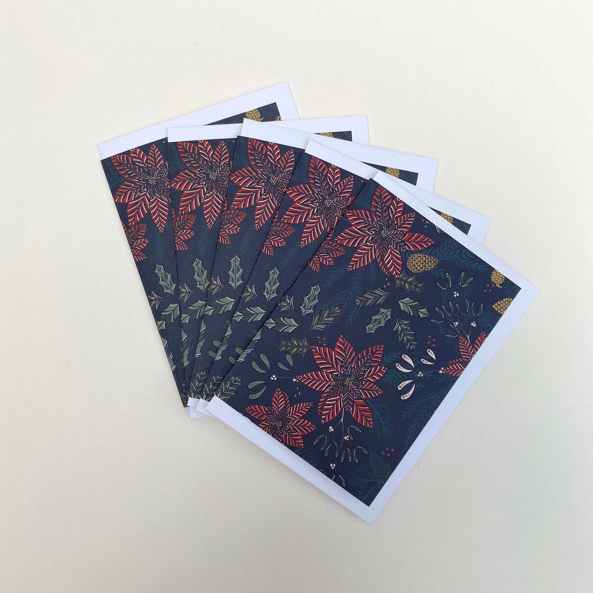A fan of rectangular Christmas  greeting cards with a dark navy blue background and a festive botanical pattern featuring red poinsettias, olive green leaves, and small gold pinecones, displayed on a light beige background.