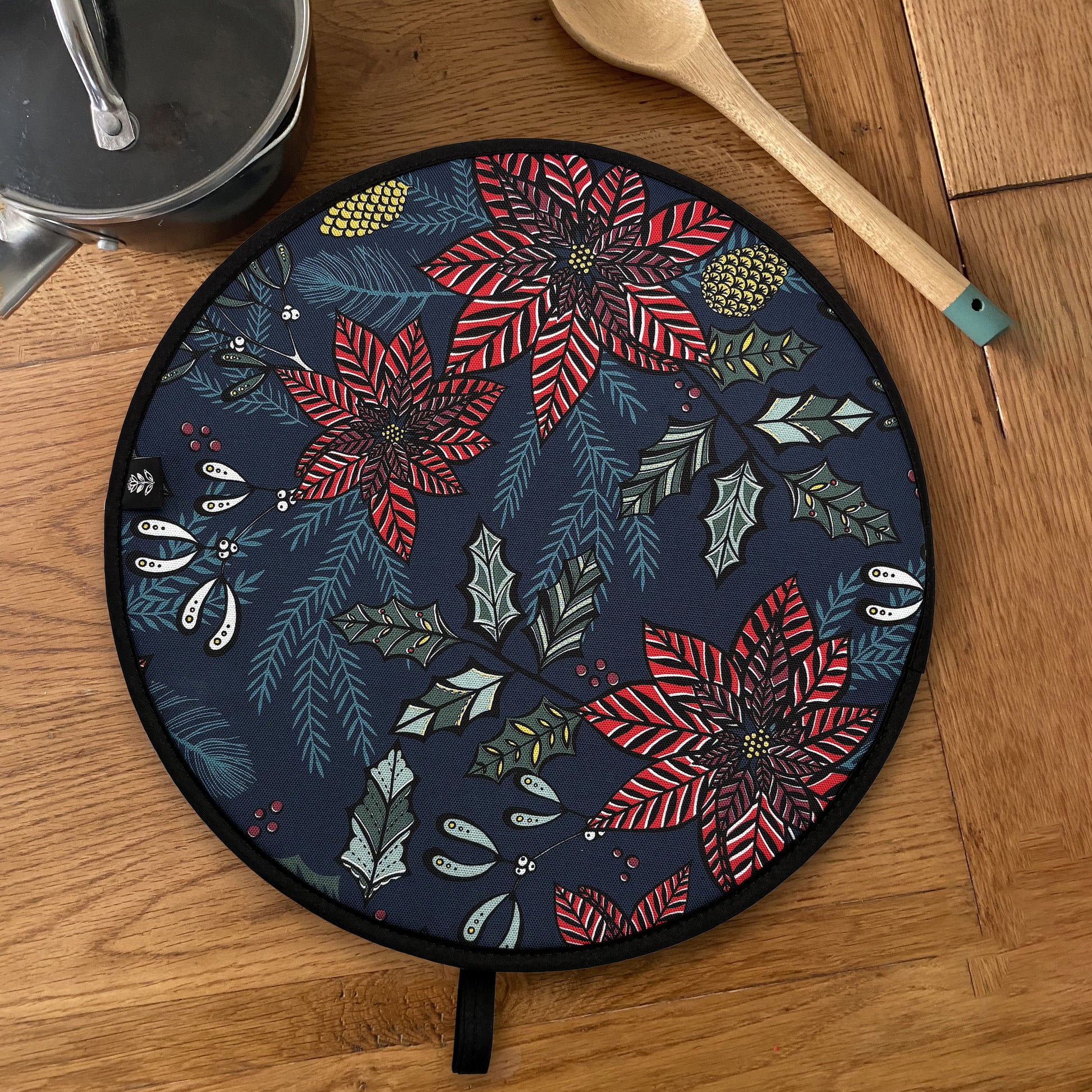 Christmas Chef pad has been placed on a wooden table.  It features red poinsettia flowers, green holly, fir, mistletoe and yellow pinecones on a navy background.