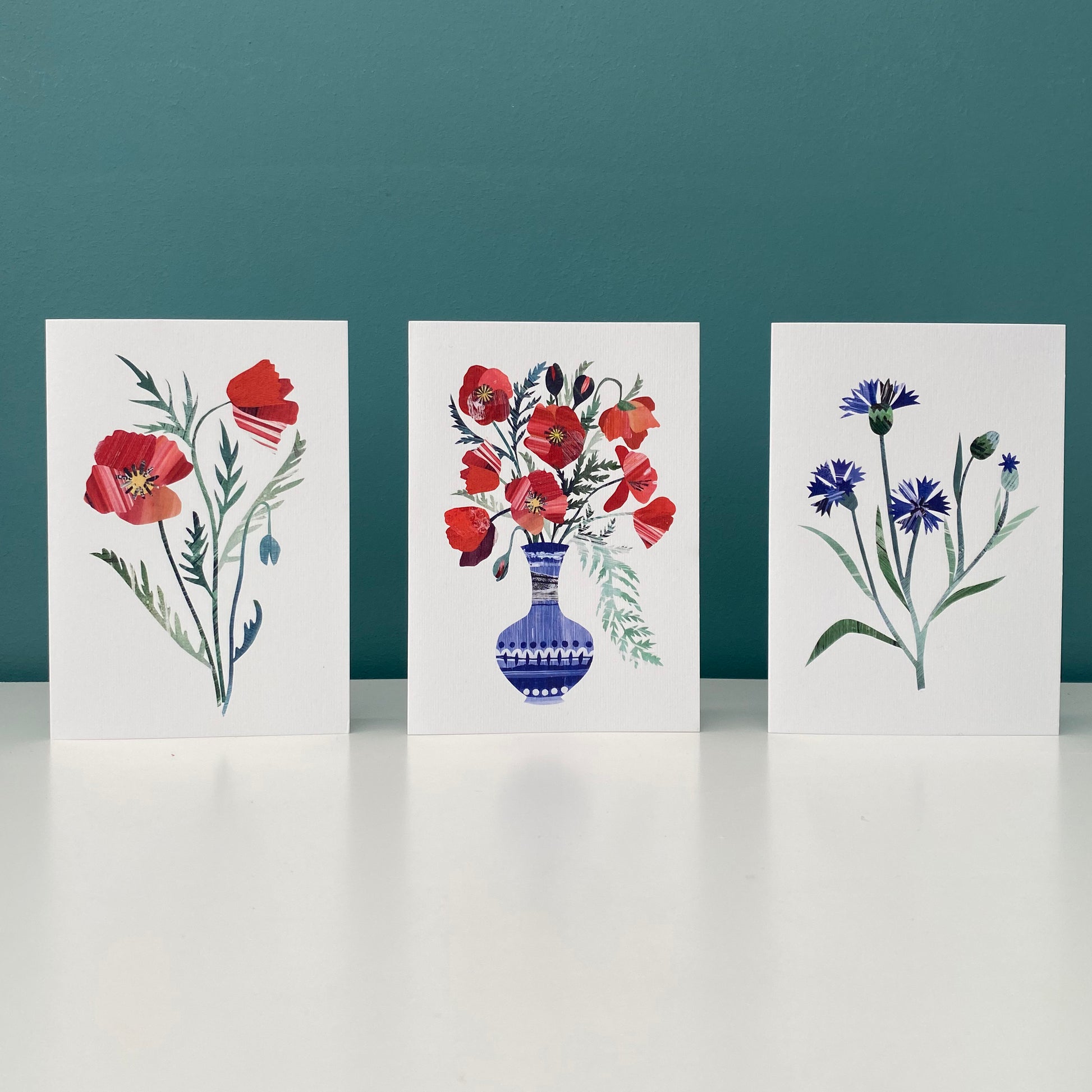 A set of 3 greeting cards featuring unique hand collaged design.  One card has a red poppy on it another Blue Corn flowers and the third hand a blue vase with red poppies in it.  They are stood on a white shelf.