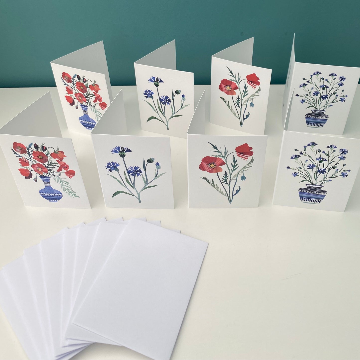 8 notecards featuring Poppies and Cornflowers are standing on a white shelf, 8 plain white envelopes are next to them.