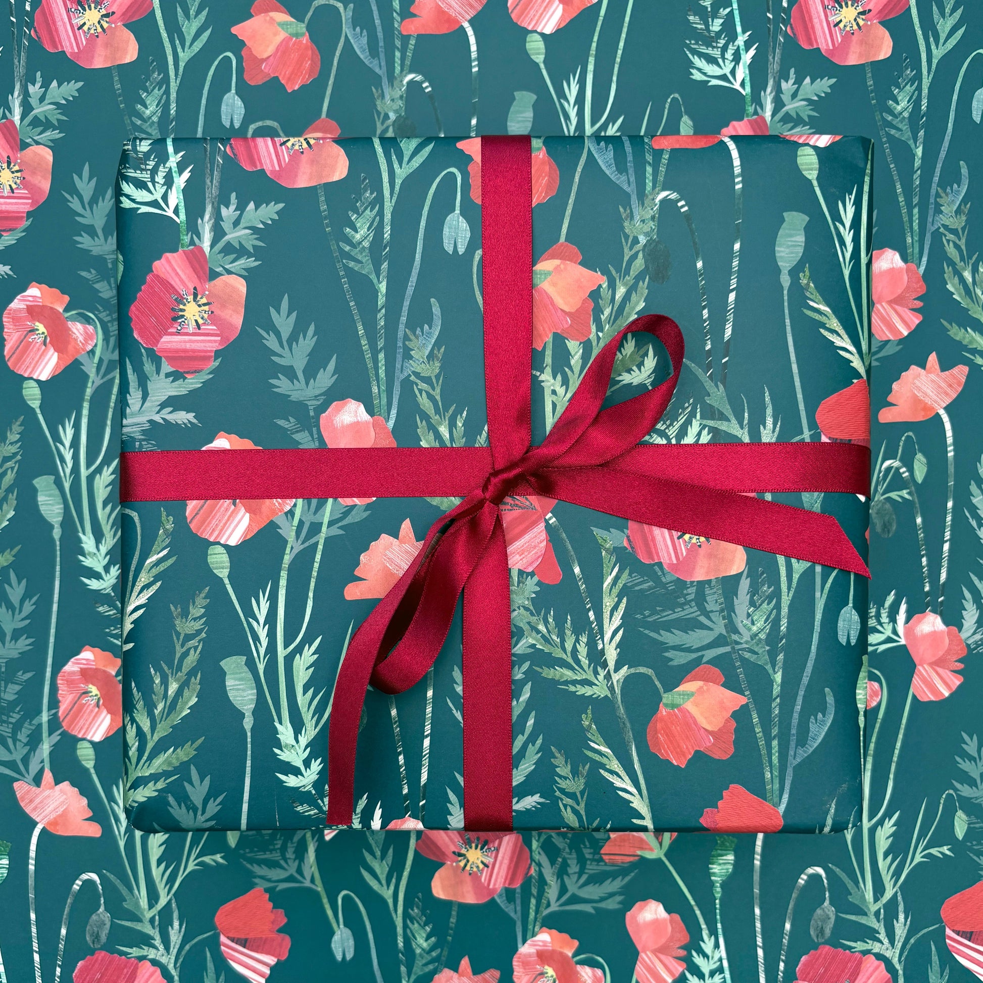 A present has been wrapped in Poppy Gift Wrap and has been tied with a coordinating Red satin ribbon and placed on a sheet of laid out Poppy  gift wrap. The gift wrap features Red Poppies with green leaves on a Dark Green background.