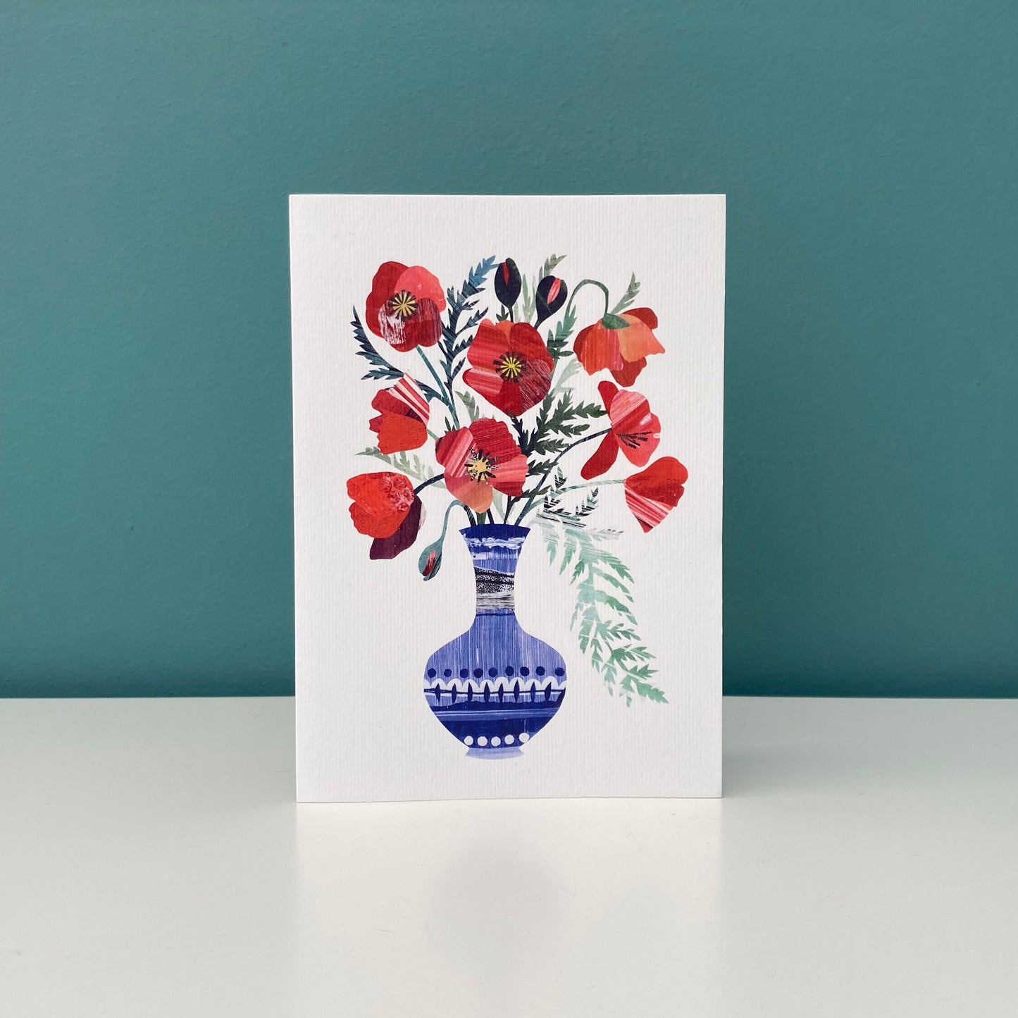 Bright Red Poppies in a blue vase greeting card featuring unique hand collaged design with Red Poppy Flowers and green leaves on a plain white background.  The card is stood up on a white shelf against a blue/green background.