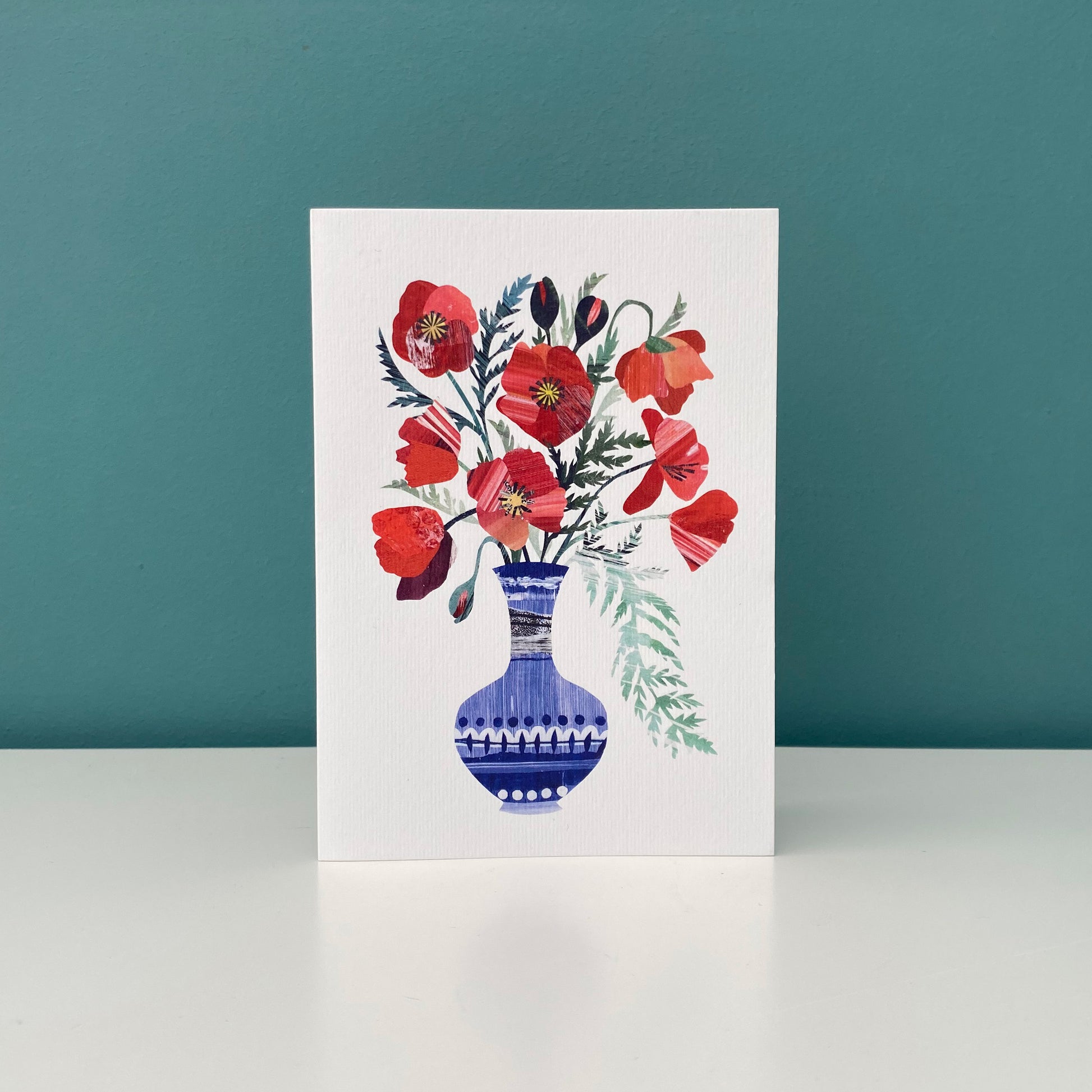 Bright Red Poppies in a blue vase greeting card featuring unique hand collaged design with Red Poppy Flowers and green leaves on a plain white background.  The card is stood up on a white shelf against a blue/green background.