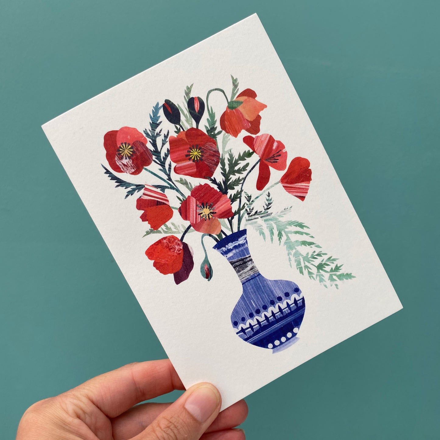 Bright Red Poppies in a blue vase greeting card featuring unique hand collaged design with Red Poppy Flowers and green leaves on a plain white background.  The card is being held against a blue/green background.