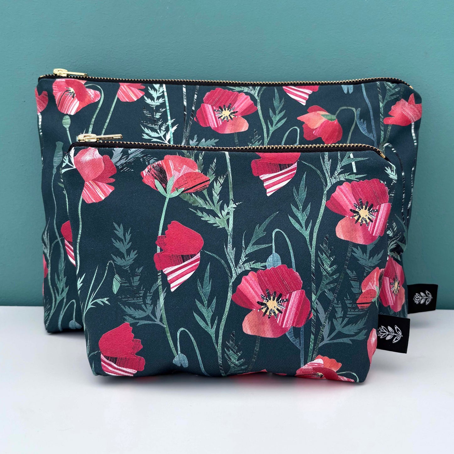 A large and small poppy wash bag have been placed on a white shelf against a blue green background.