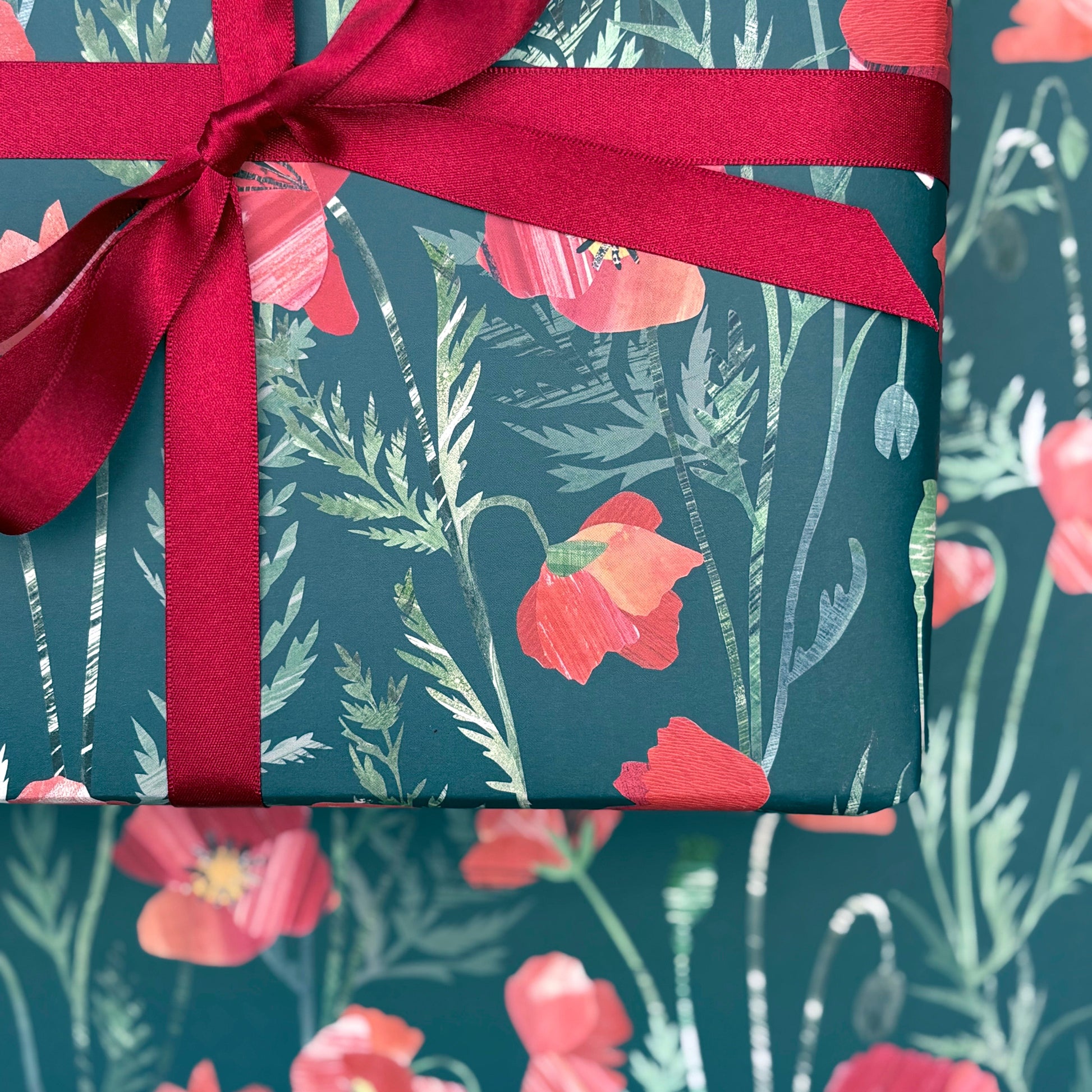 A close up of a present that has been wrapped in Poppy Gift Wrap and tied with a coordinating red satin ribbon and placed on a sheet of laid out Poppy gift wrap.