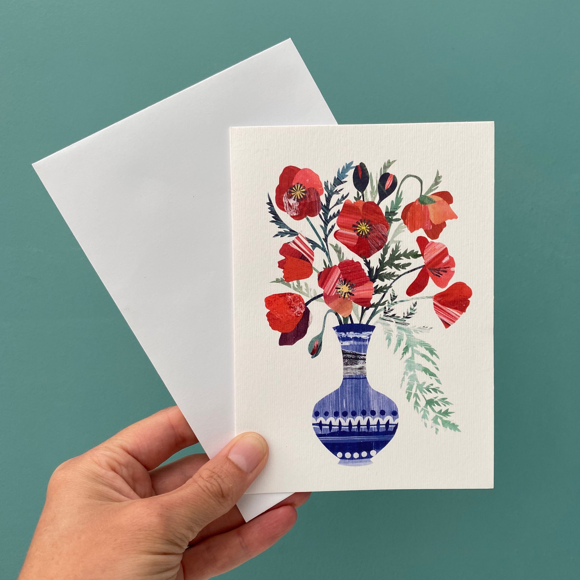 Bright Red Poppies in a blue vase greeting card featuring unique hand collaged design with Red Poppy Flowers and green leaves on a plain white background.  The card and a white envelope is being held up against a blue/green background.