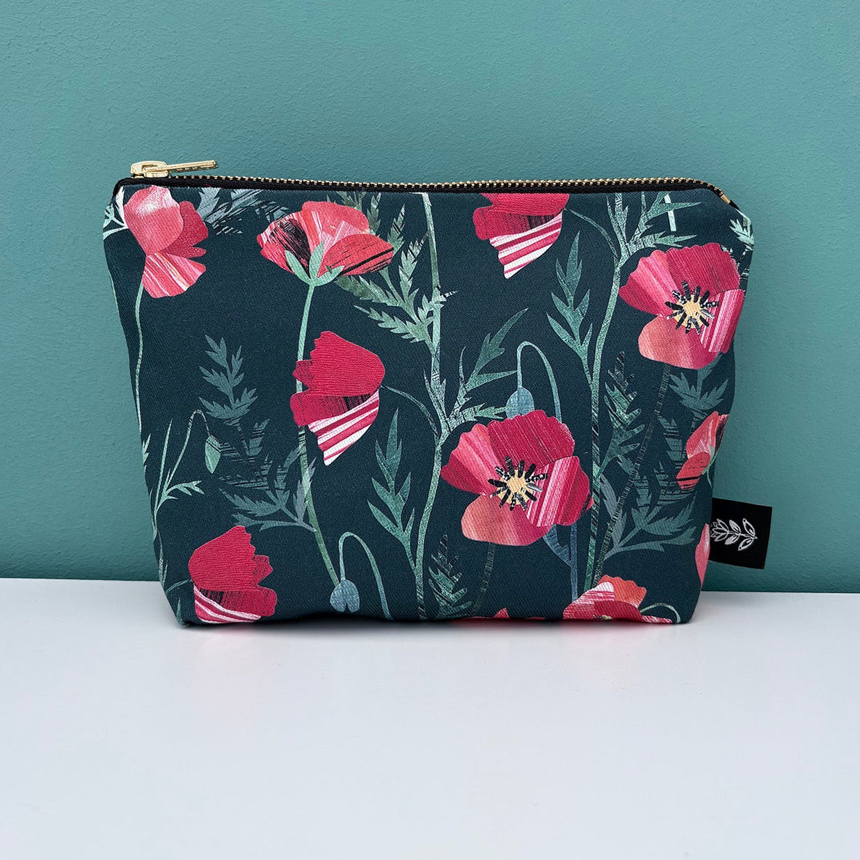A small Poppy make up bag has been placed on a white shelf against a blue green wall.
