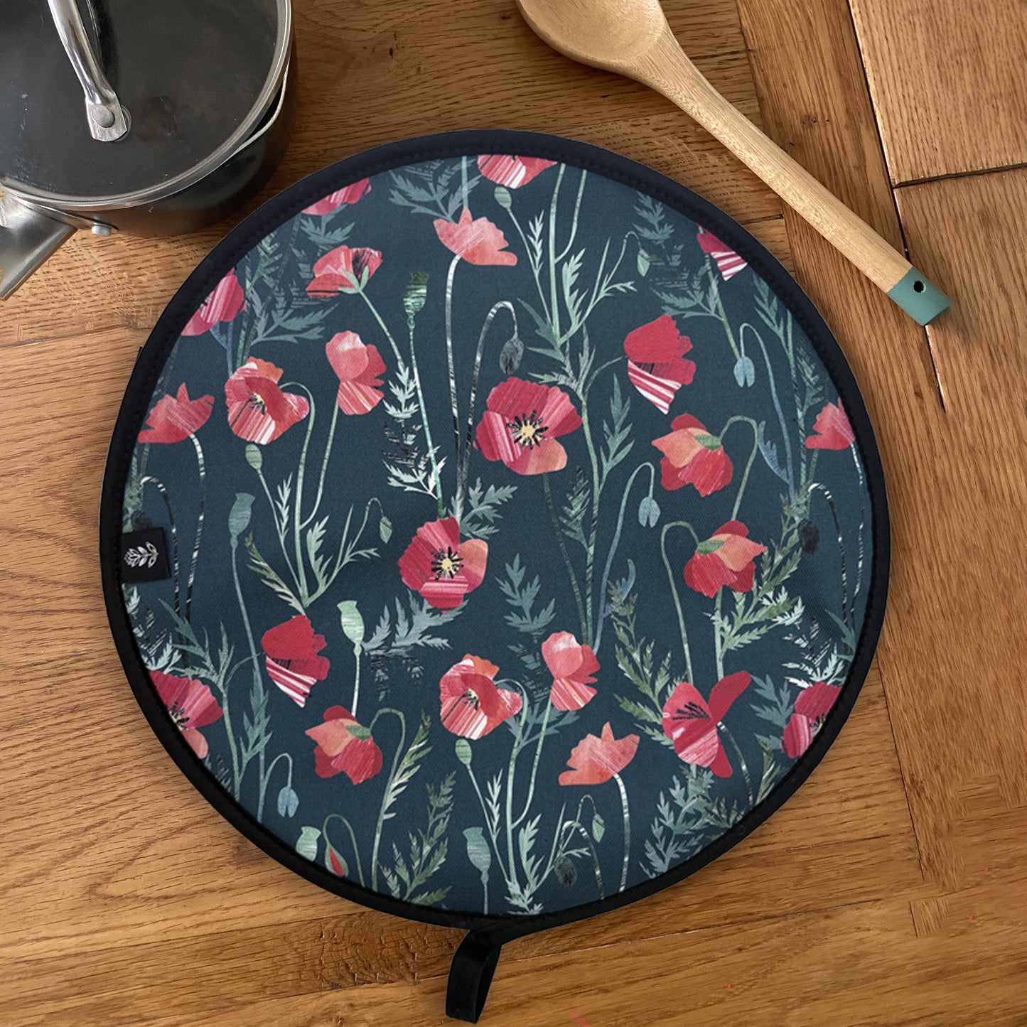 A Poppy Aga Cover has been placed on a wooden table with a saucepan and a wooden spoon. The poppy pattern features pretty red poppy flowers on a dark green background.
